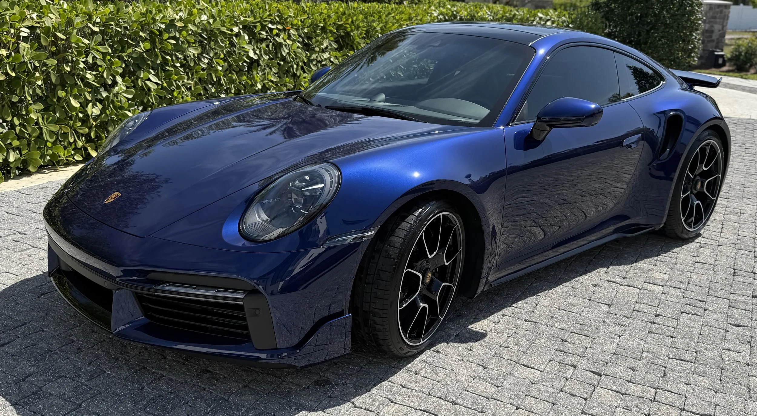 Blue Porsche sports car parked on a cobblestone driveway beside green bushes.