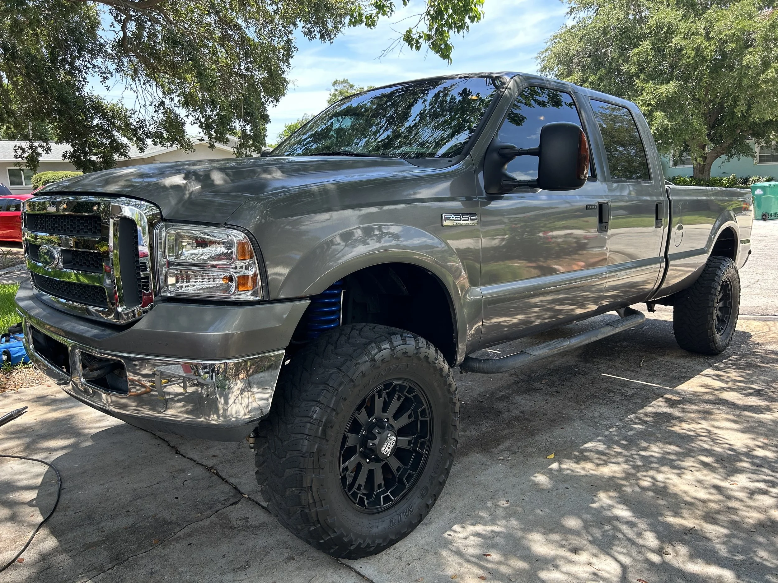 A gray Ford F-350 Super Duty pickup truck with black wheels, off-road tires, and tinted windows parked on a driveway under a tree with green leaves.