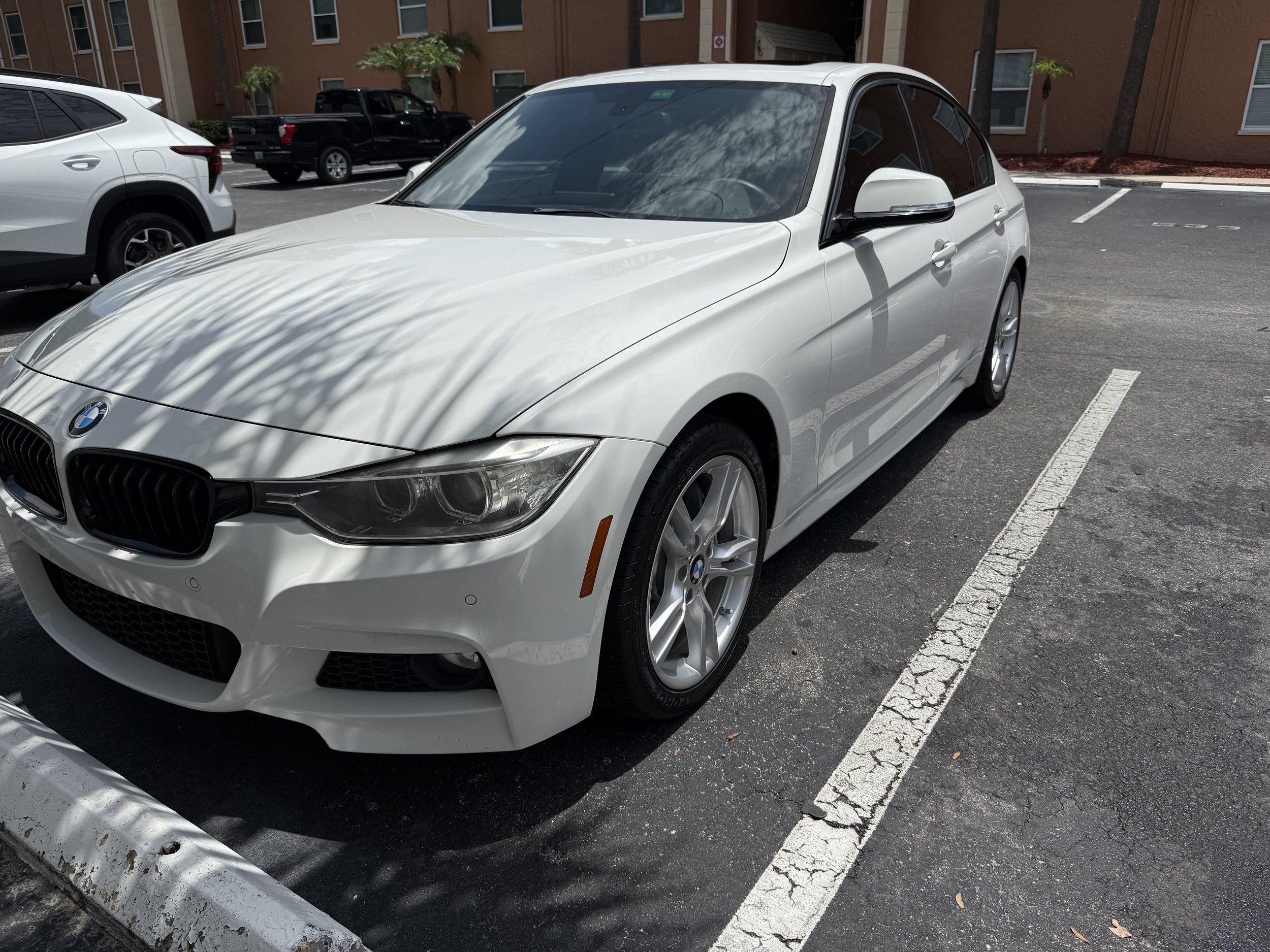 A white BMW coupe parked in a parking lot with other vehicles and a red brick building in the background.