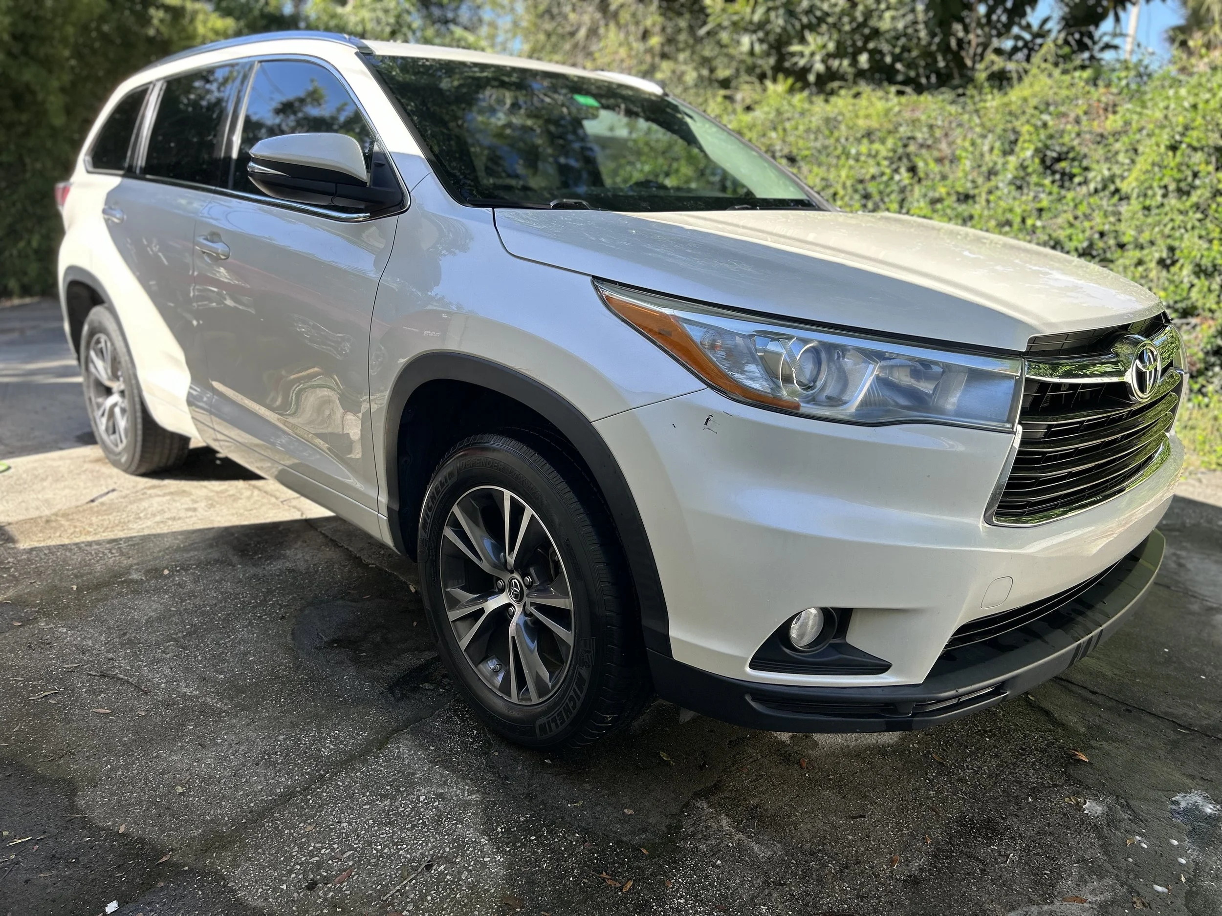 White Toyota SUV parked on a paved surface near green bushes, with sunlight reflecting off its surface.