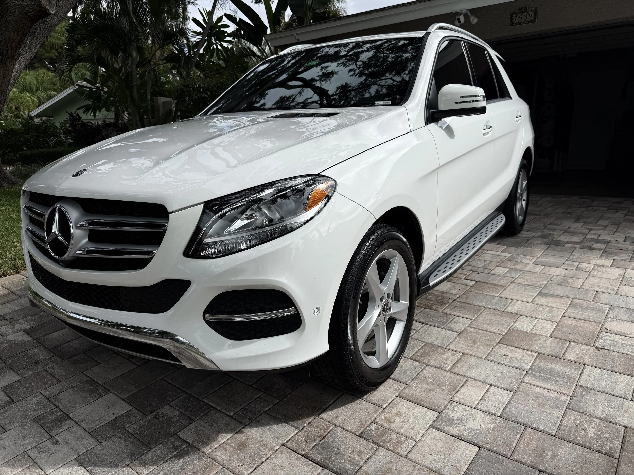 White Mercedes-Benz SUV parked on a brick driveway with trees and house in the background.