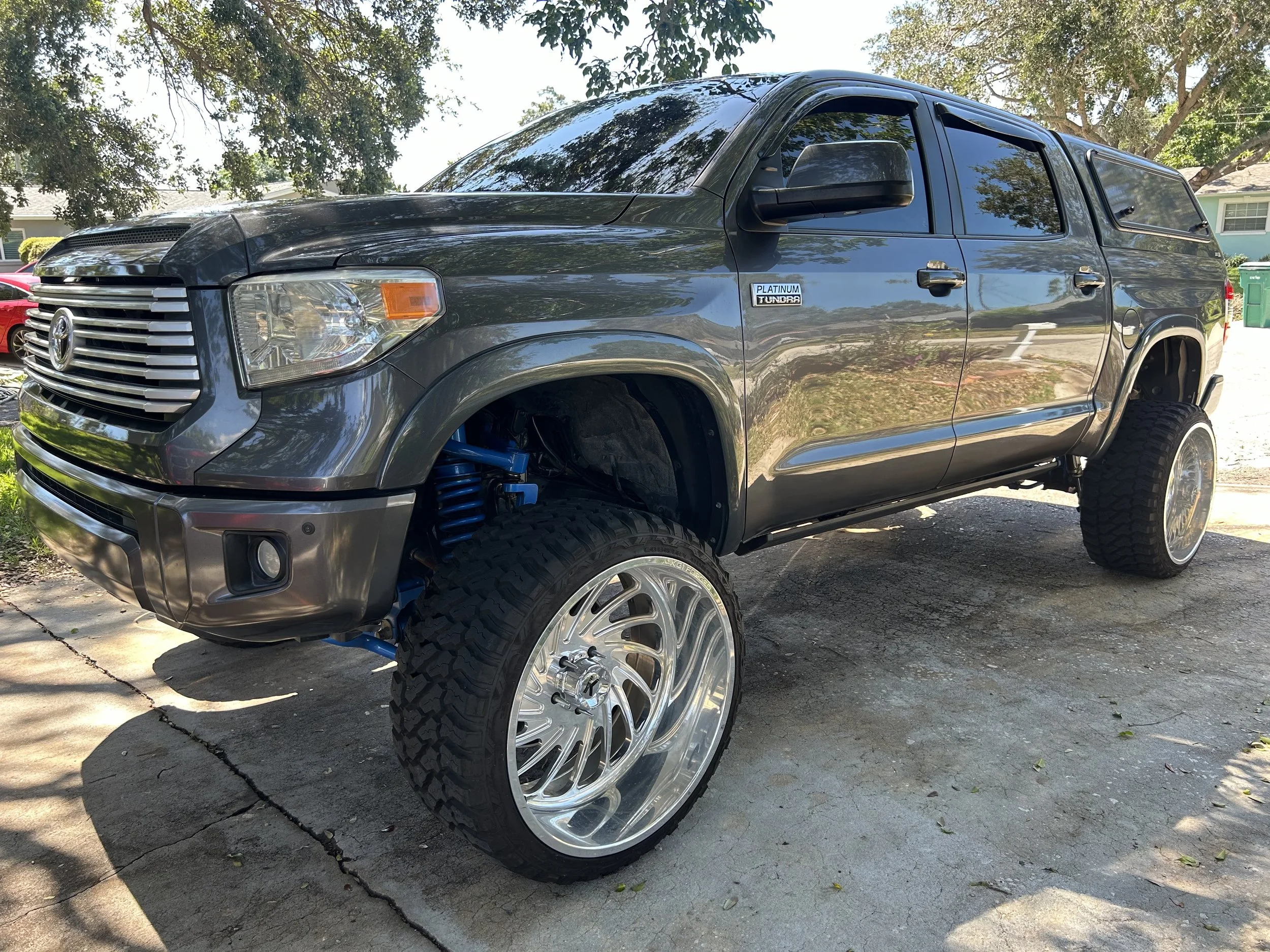 A black Toyota Tacoma pickup truck with large custom wheels, lifted suspension, and a canopy on the back, parked on a driveway under tree shade.