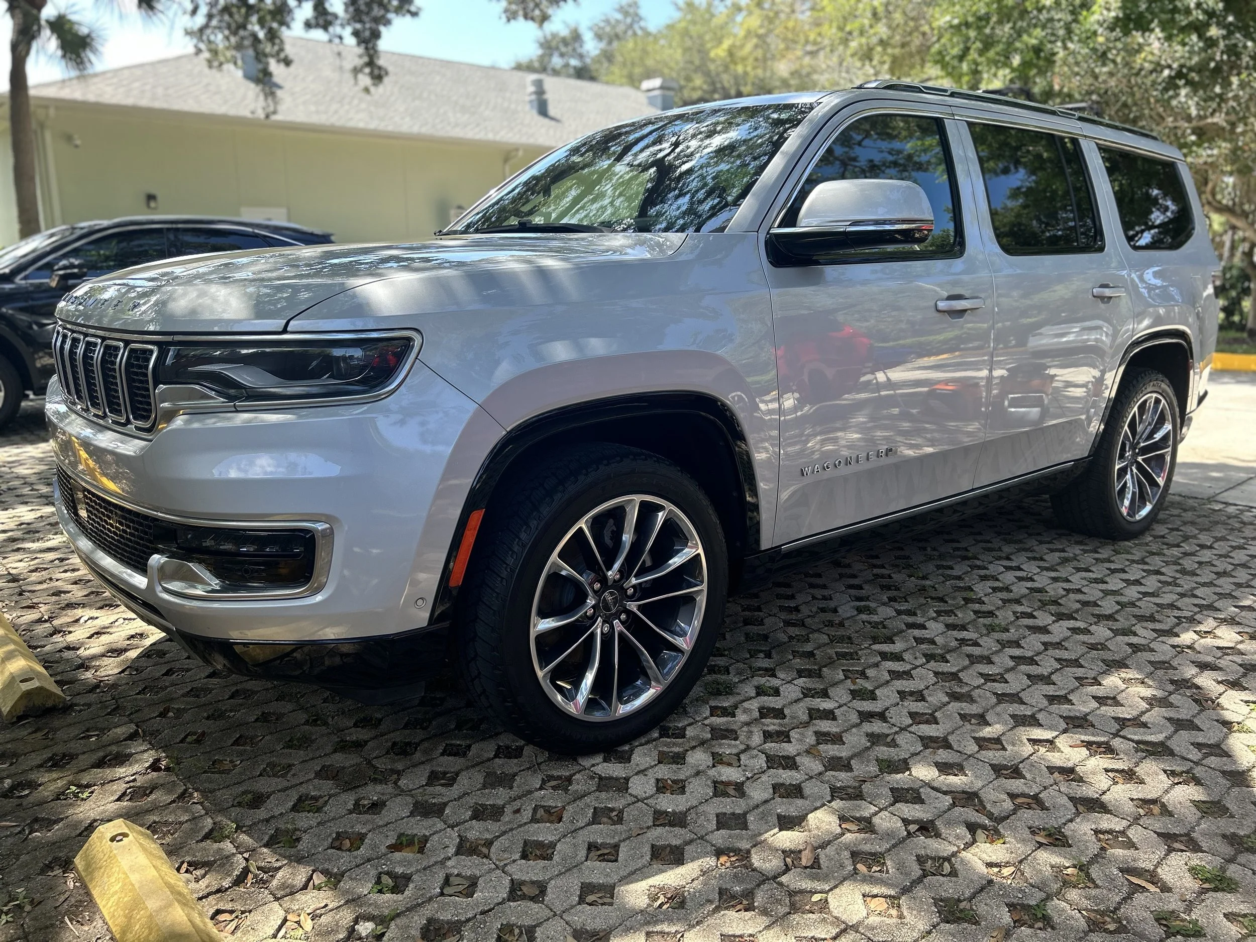 Silver Jeep Wagoneer parked on a cobblestone parking lot with trees and a house in the background.