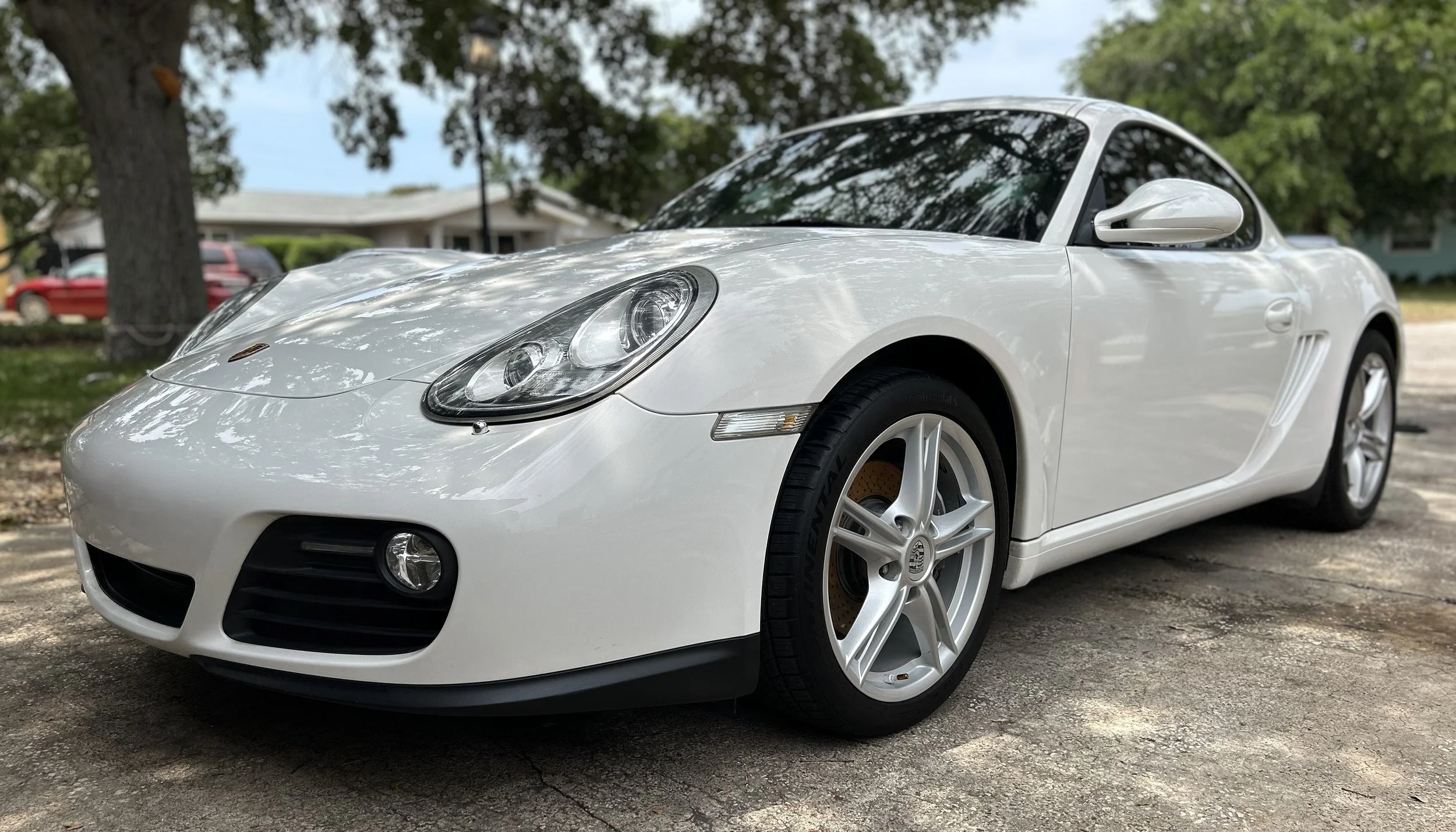 A white Porsche sports car parked on a driveway with residential houses and trees in the background.
