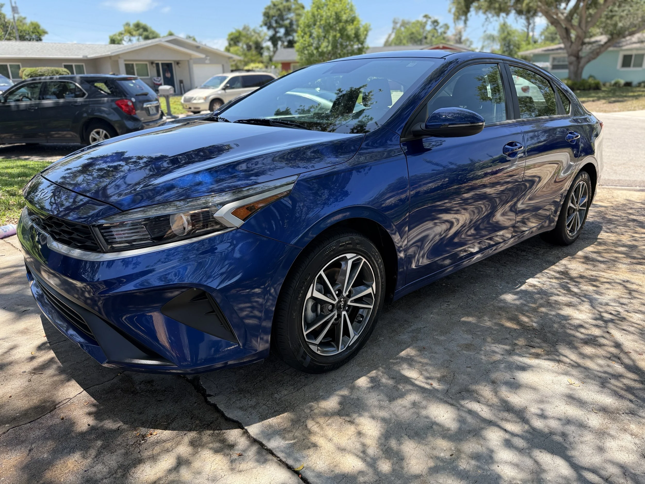 A blue sedan parked on a driveway with a house and other parked cars in the background. The car has a sleek design with black and silver alloy wheels and reflects the surrounding trees and sky on its surface.