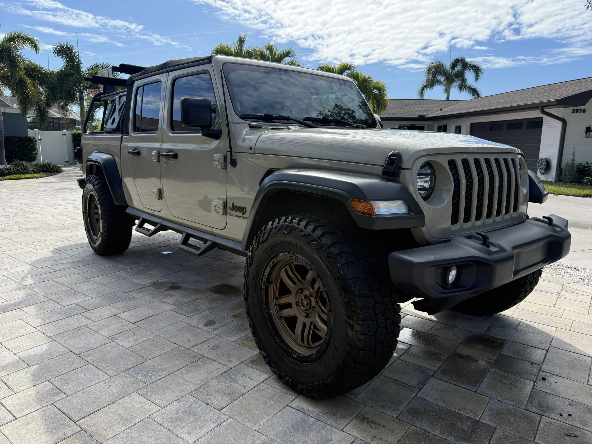 A beige Jeep Gladiator Rubicon parked on a paved driveway in front of a suburban house with palm trees, a blue sky, and some clouds in the background.