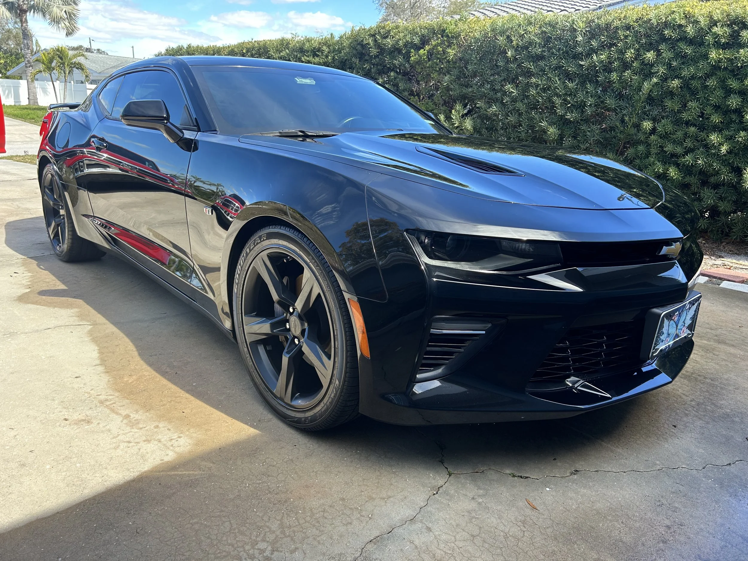 Black Chevrolet sports car parked on concrete pavement with greenery in the background under a partly cloudy sky.