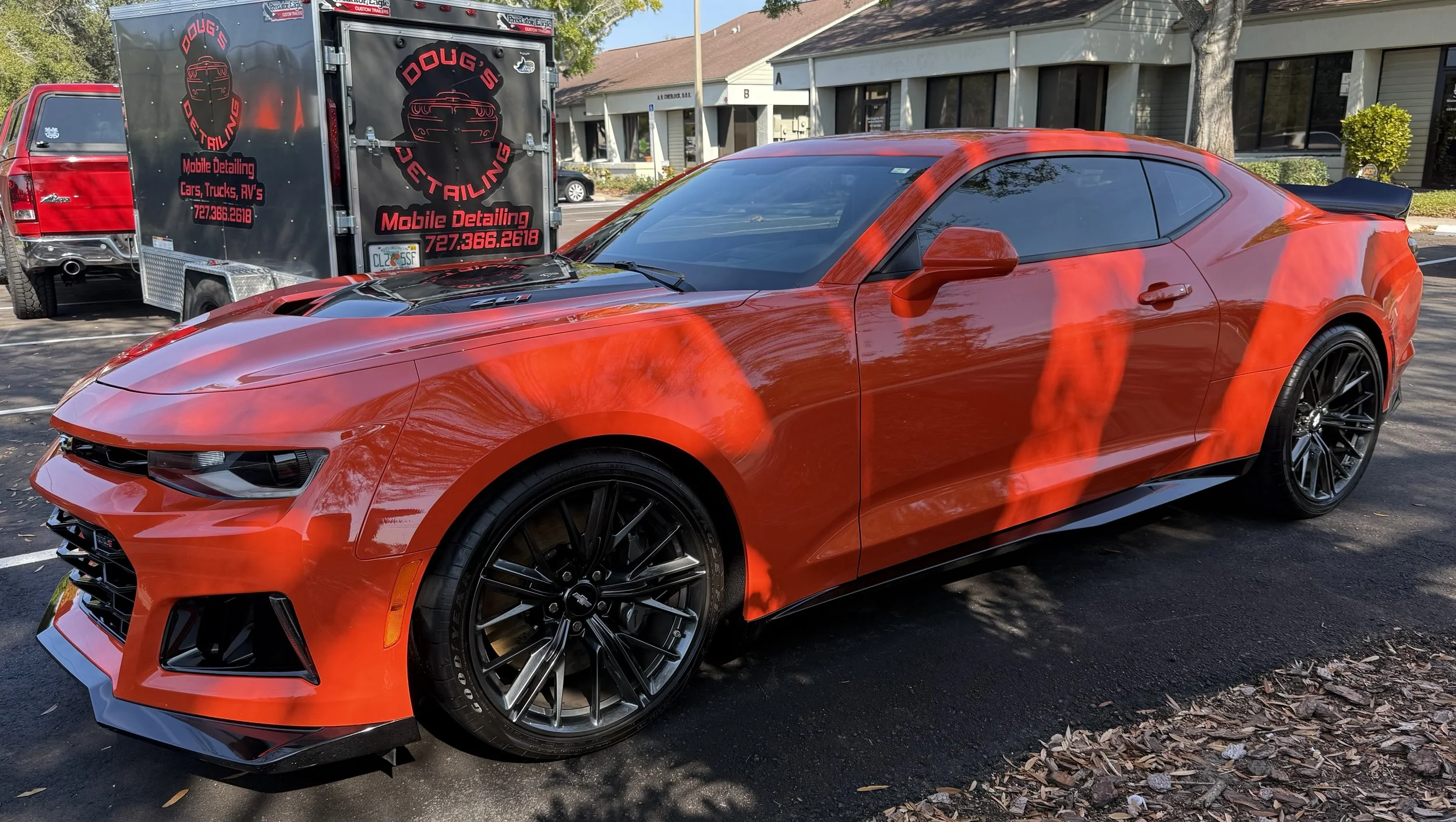 A red sports car parked on the street, with black rims and tinted windows, alongside a black trailer for mobile detailing services.