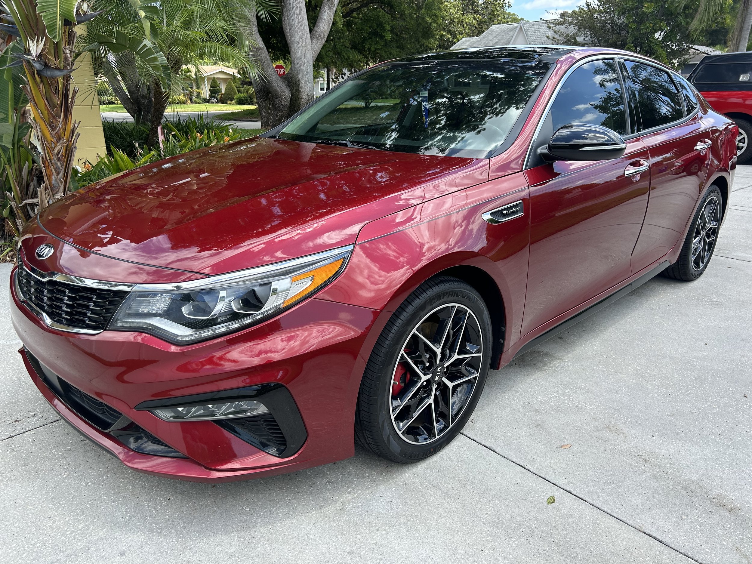 A red Kia sedan parked on a concrete driveway with trees and houses in the background.