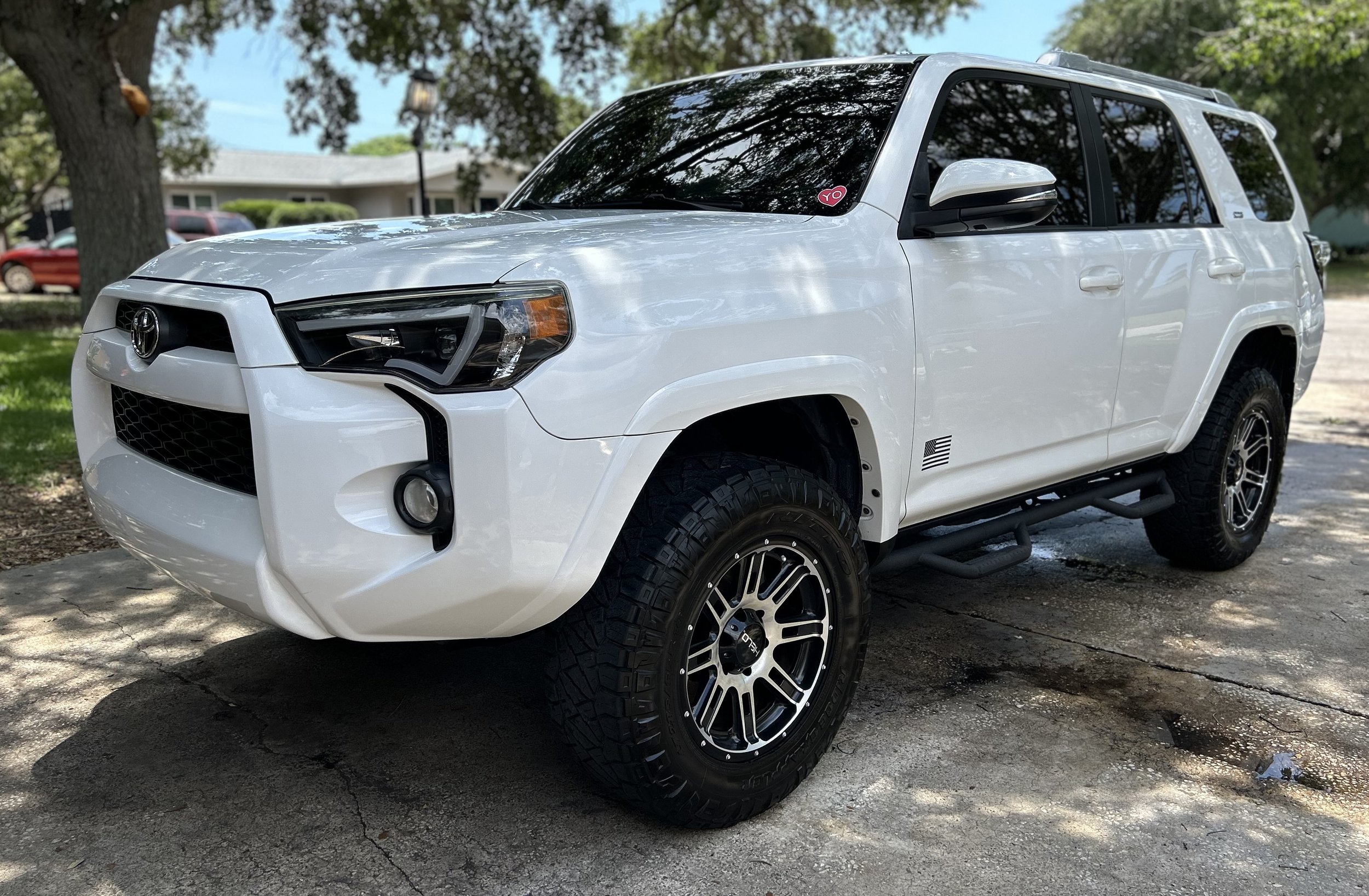 White Toyota 4Runner SUV parked on a driveway under a tree with green leaves and a house in the background.