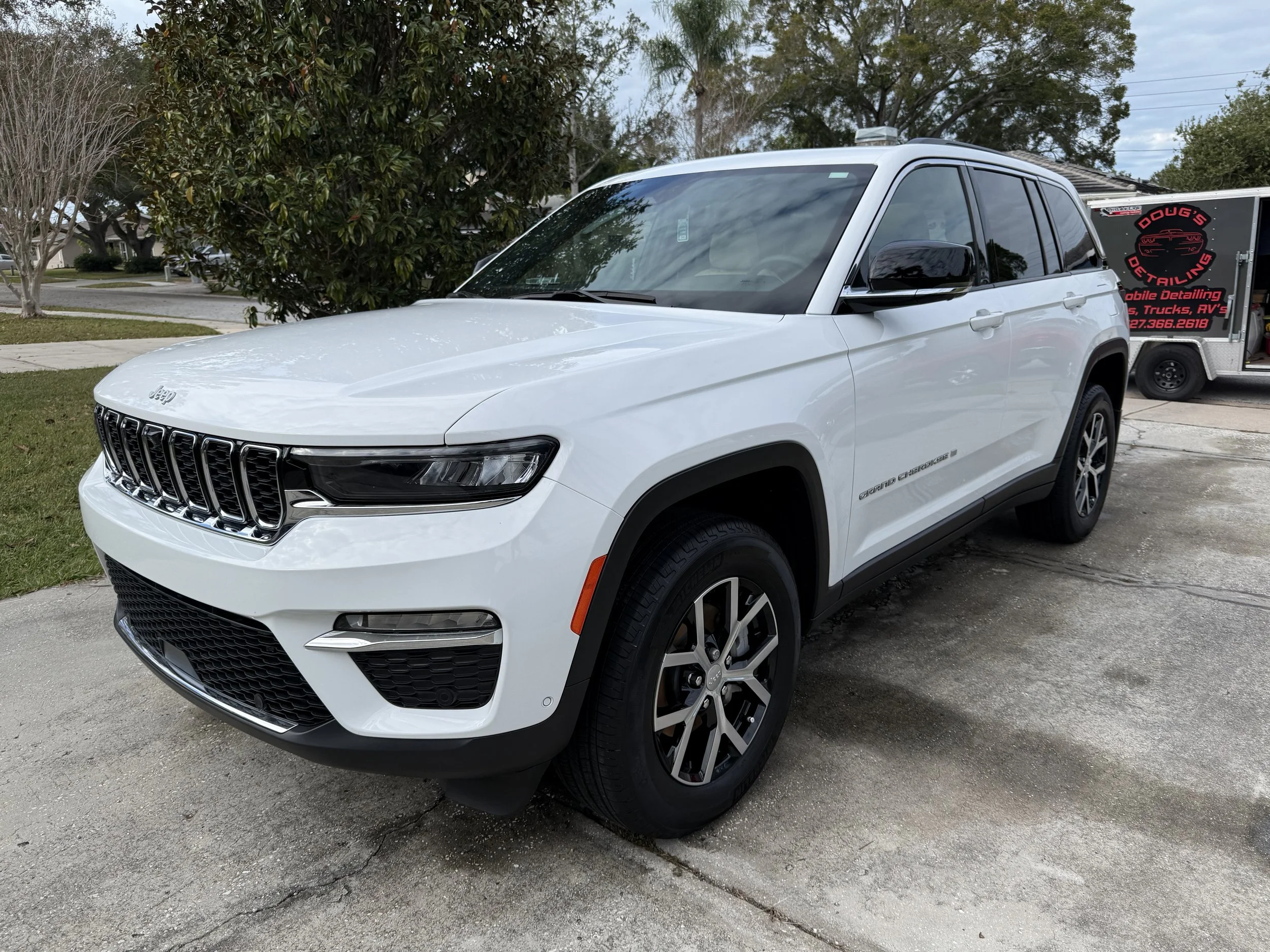 White Jeep Grand Cherokee parked on driveway with trees and a mobile detailing truck in the background.