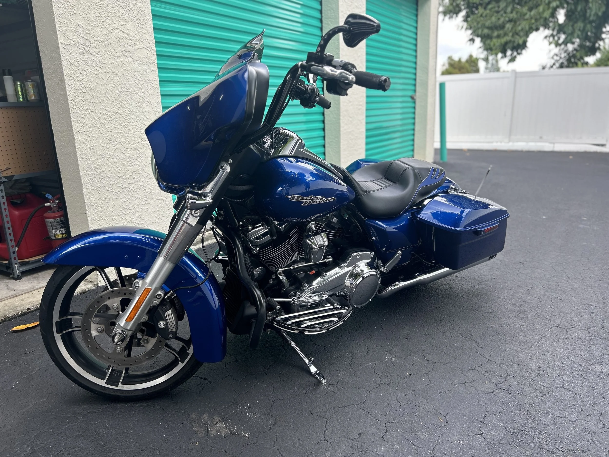 Blue Harley Davidson touring motorcycle parked on black asphalt near a beige textured wall with a teal rolling shutter and white fence in the background.