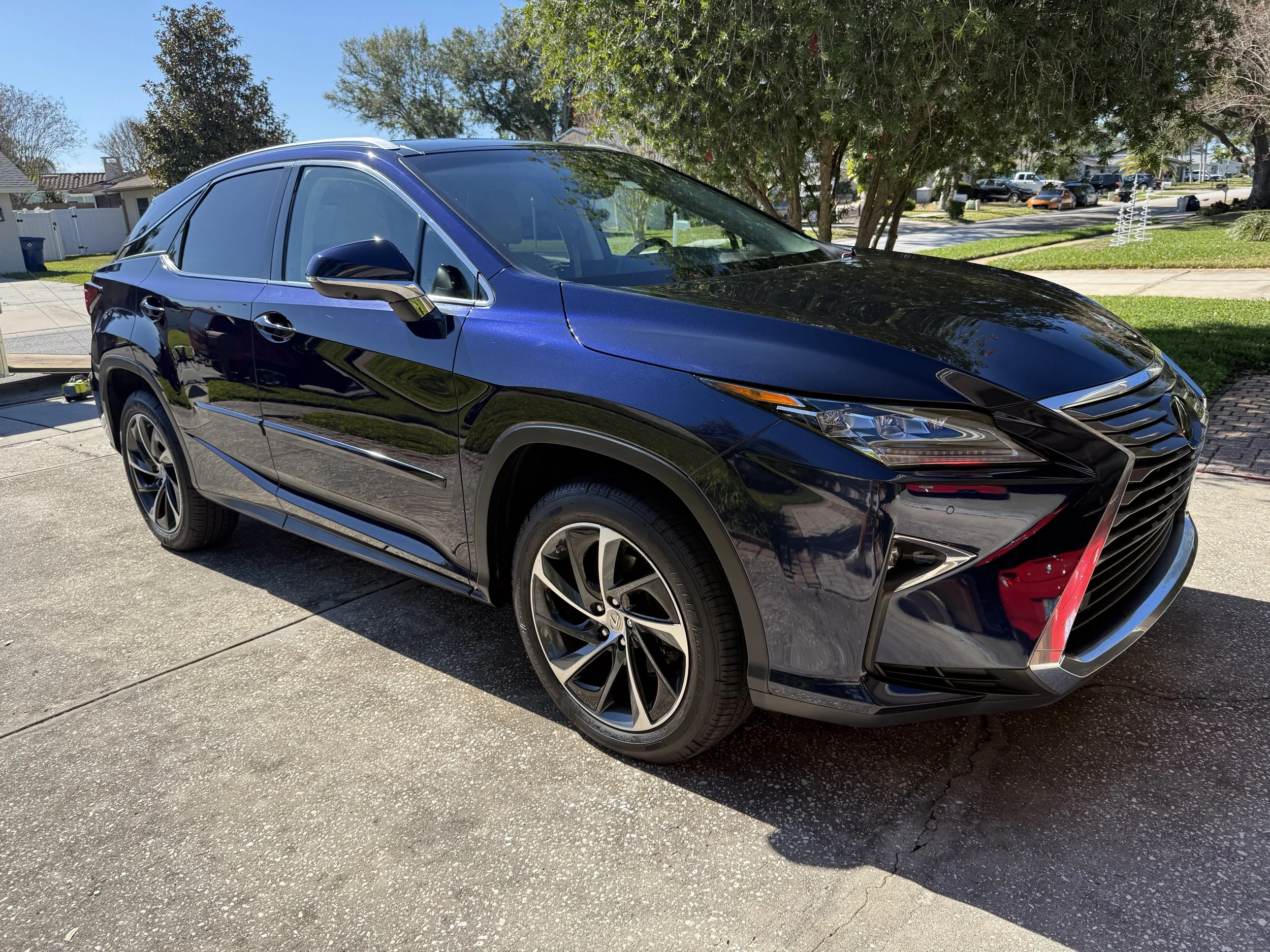 A shiny black Lexus RX parked on a concrete driveway on a sunny day, with trees and parked cars in the background.