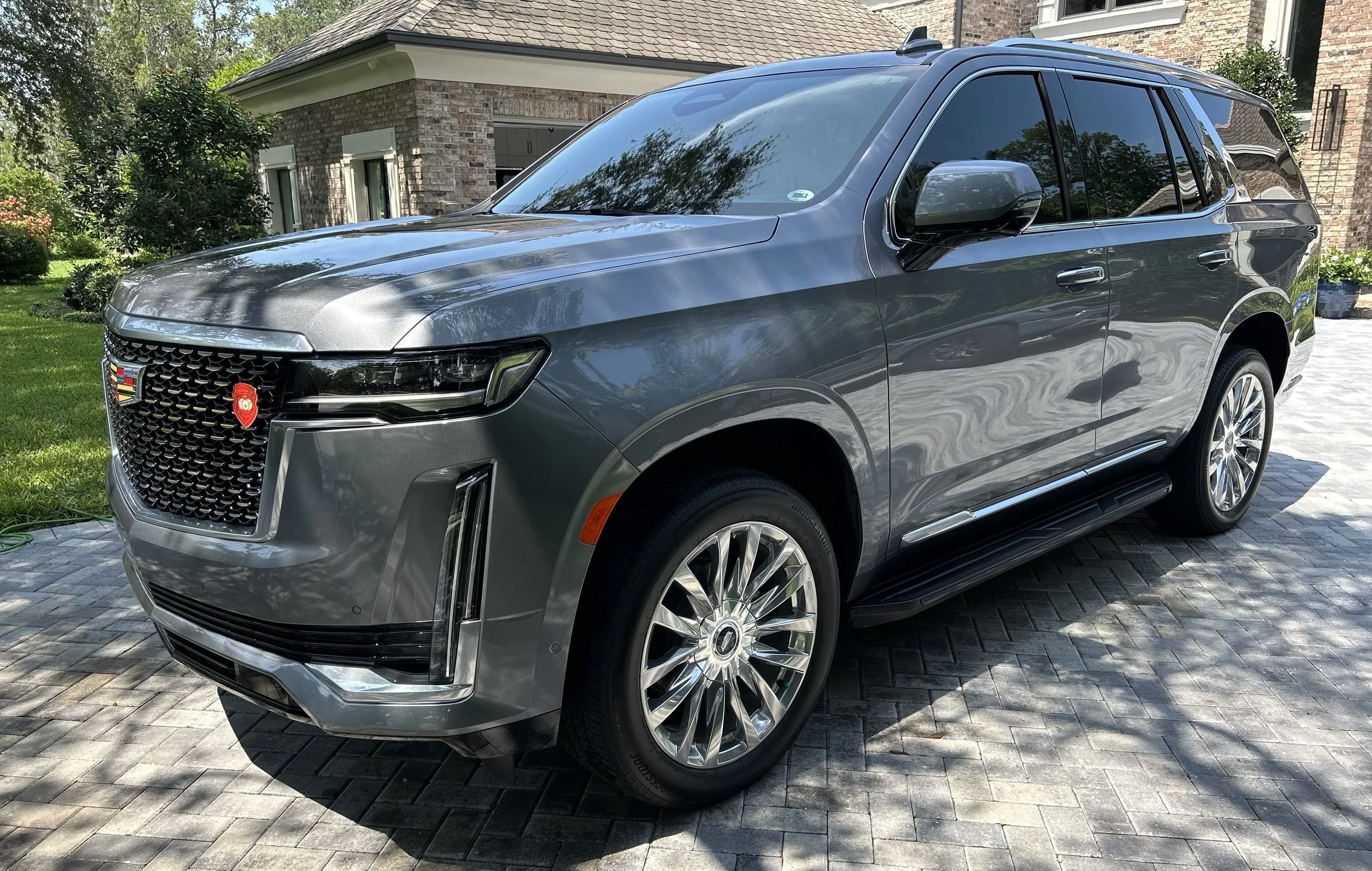 A gray Cadillac SUV parked on a driveway with a brick house and greenery in the background.