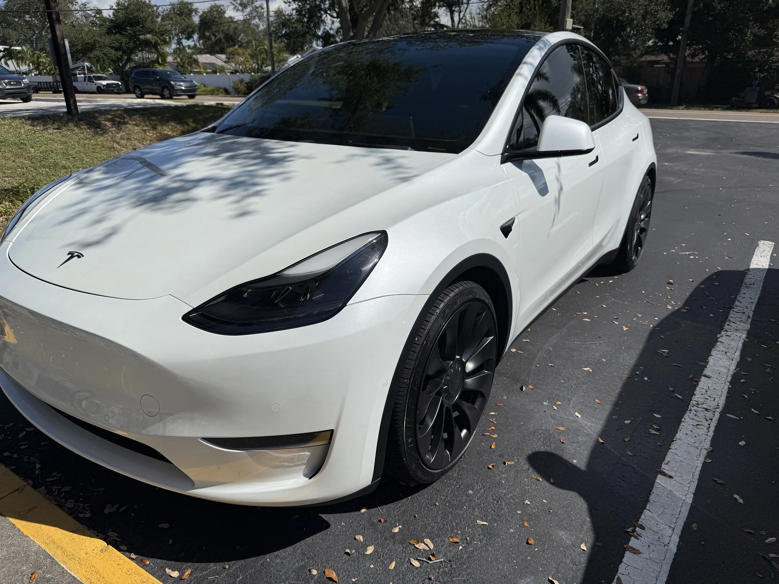 White Tesla Model 3 parked in a parking space with trees and other cars in the background.