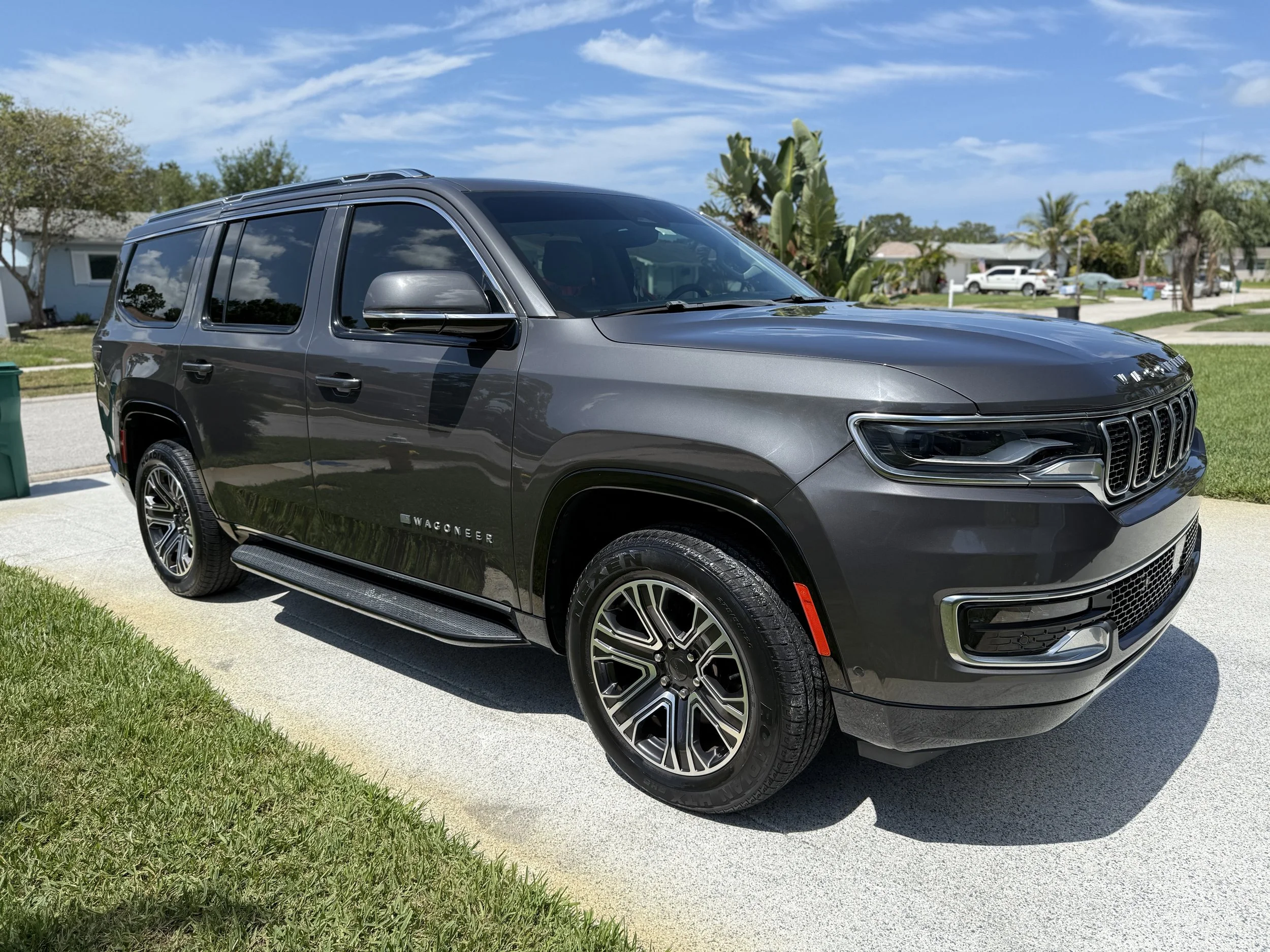 A gray Jeep Wagoneer SUV parked on a driveway in a residential neighborhood on a sunny day.