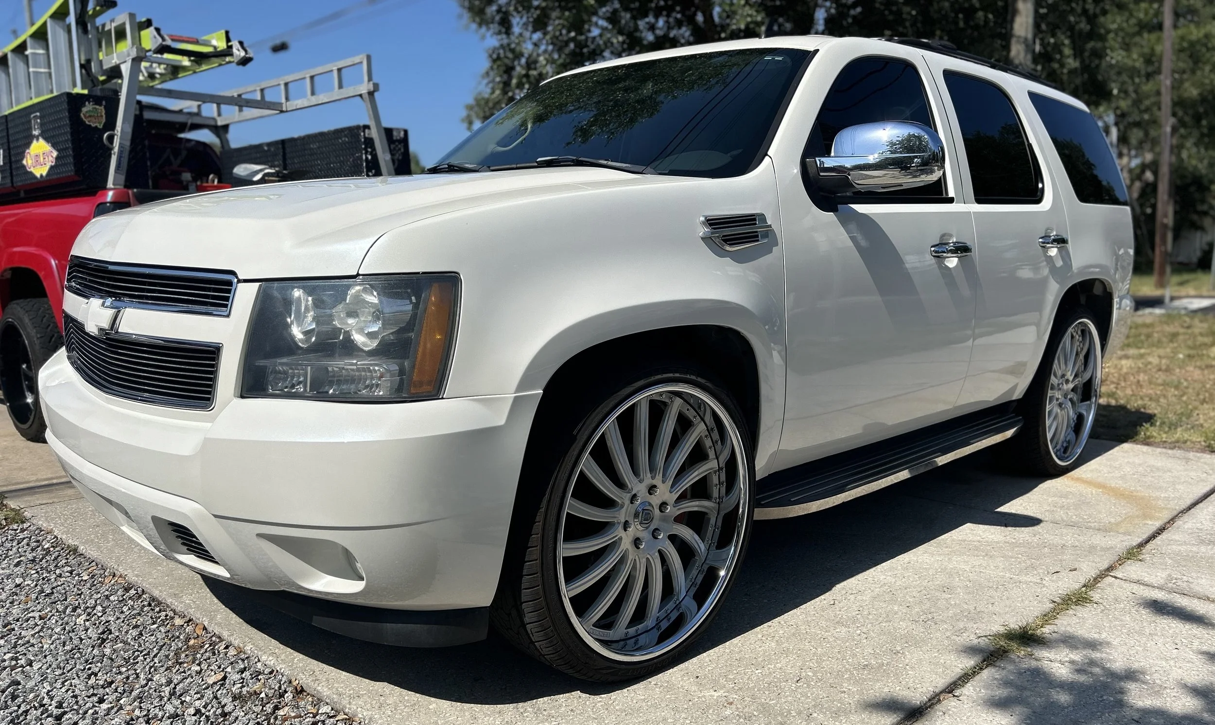 White SUV with aftermarket large rims parked on a driveway, with a red truck and equipment in the background.