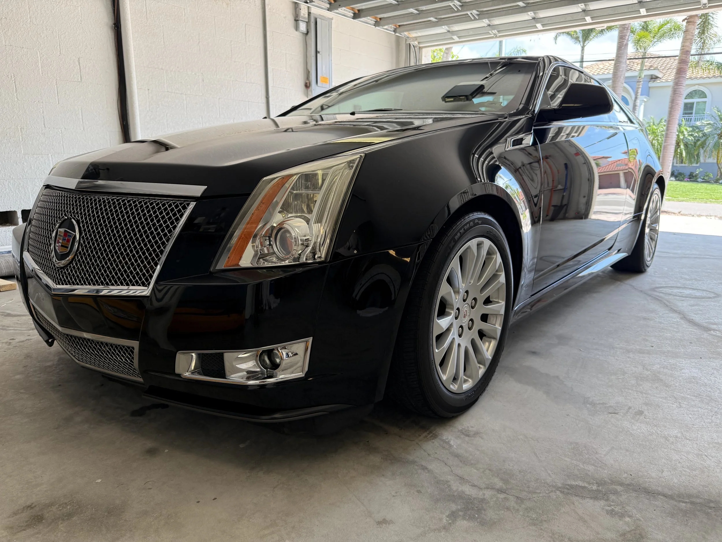 Black Cadillac CTS coupe parked inside a garage with a partial view of a suburban neighborhood outside.