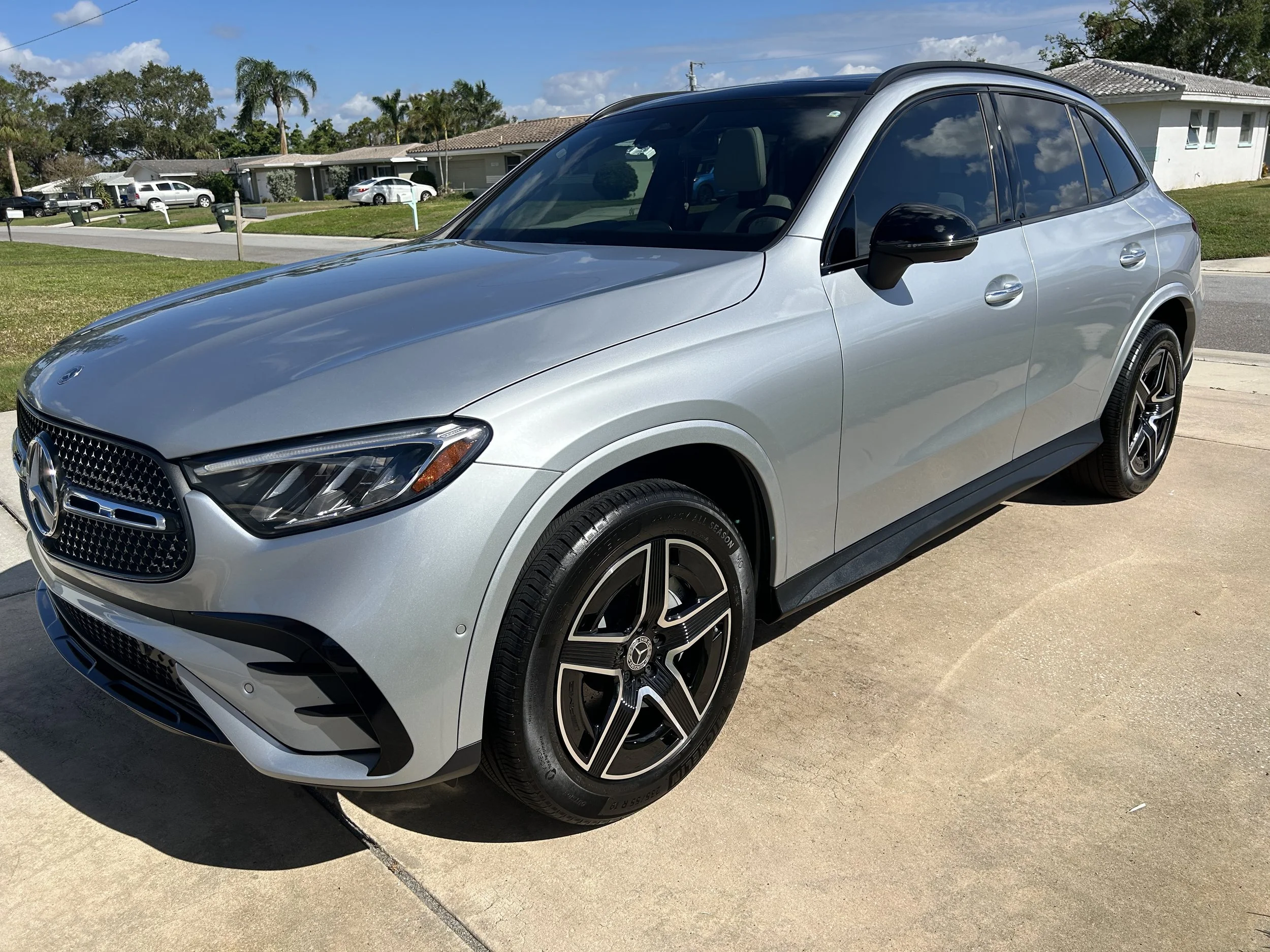 Silver Mercedes-Benz SUV parked on a driveway in front of a residential neighborhood with houses, green lawns, and trees under a partly cloudy sky.
