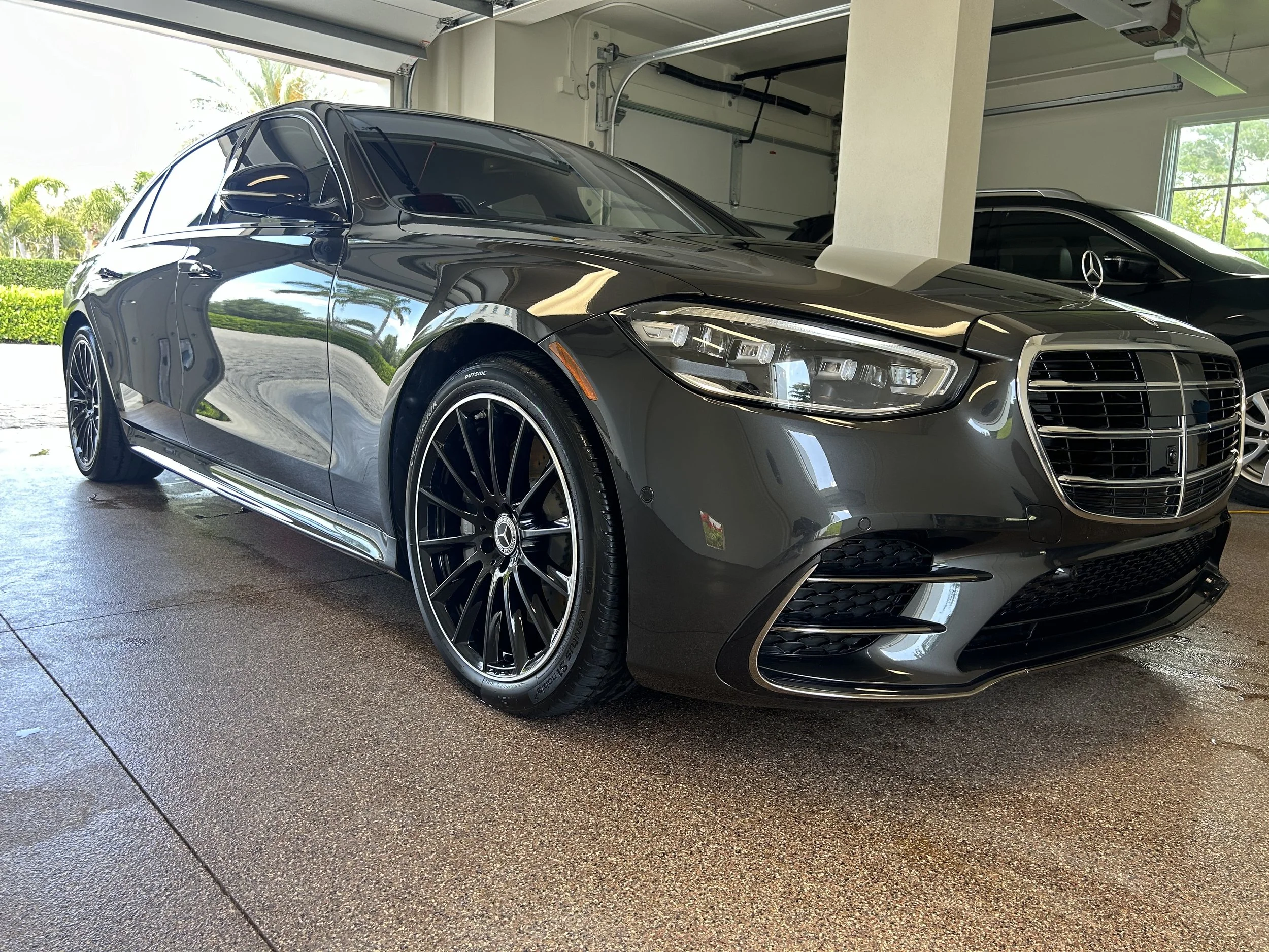 Black Mercedes-Benz sedan parked inside a garage with other cars, with windows showing green trees outside.