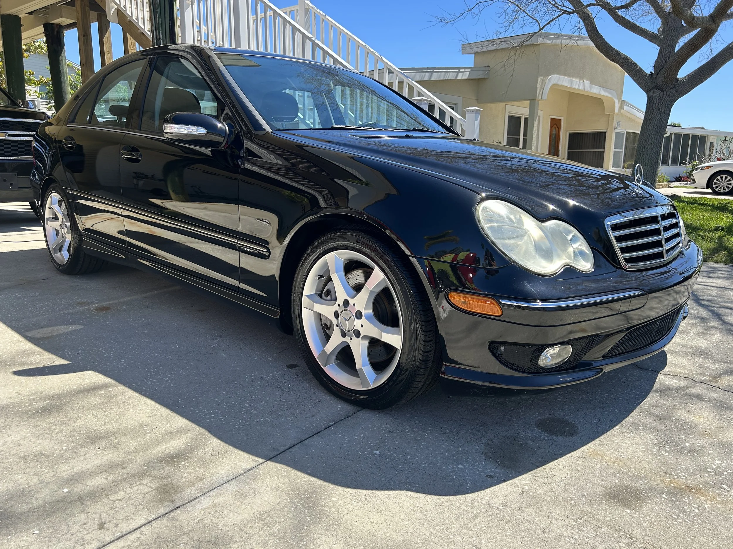 Black Mercedes-Benz sedan parked on concrete driveway in front of residential house with front yard, tree, and neighboring houses visible.