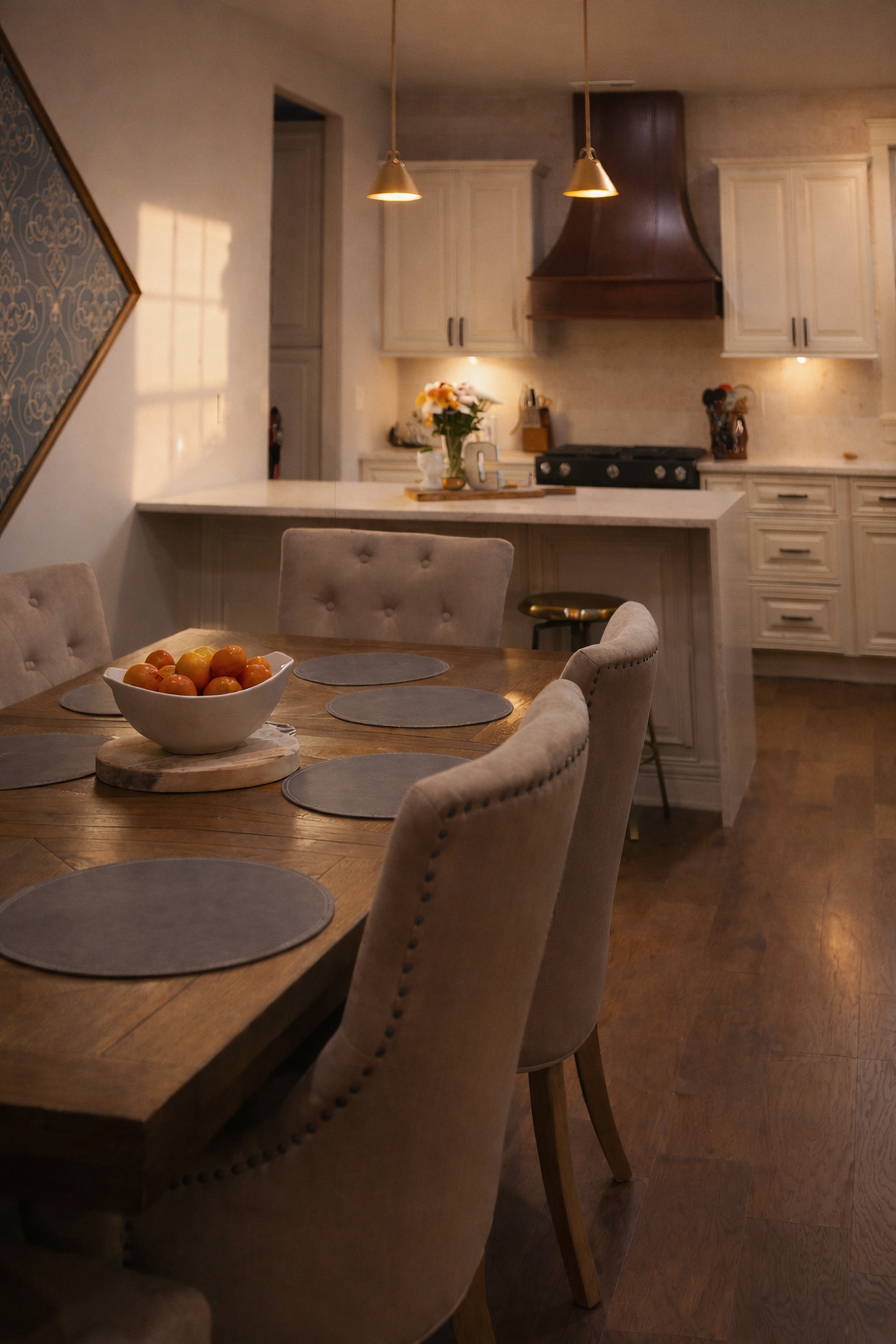 A cozy kitchen and dining area with a wooden table, beige upholstered chairs, a bowl of apples, a vase of flowers, and warm lighting.