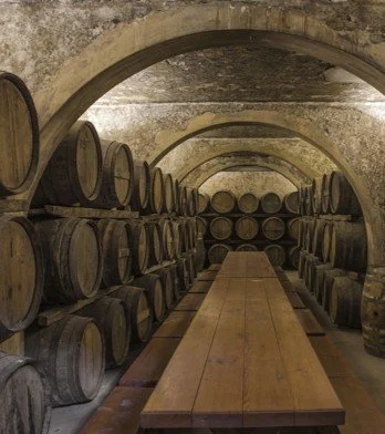 An underground wine cellar with rows of stacked wine barrels along the walls, and a long wooden table in the center.