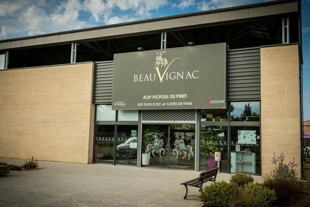 A building with a large sign reading 'Beau Vignac' and featuring a leafless tree logo. The signage indicates it is related to wine production in Pinet, France, with additional text about local wine regions. The building has glass doors with decorative horse and rider artwork and is surrounded by a paved area and some plants.