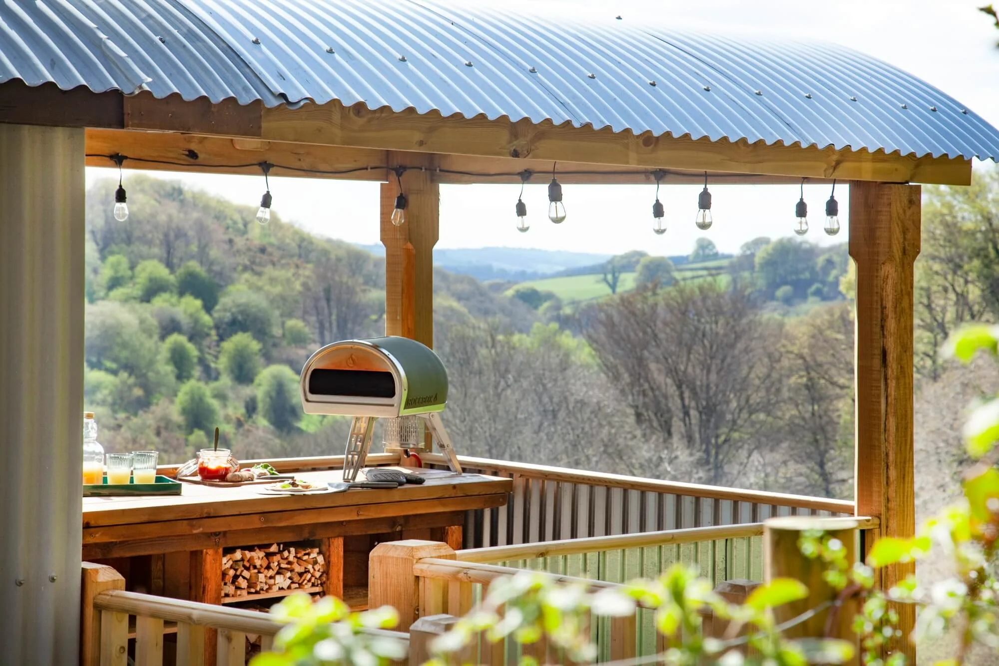 A backyard outdoor kitchen with a brick counter, wood logs stored underneath, and a pizza oven, overlooking a scenic landscape of rolling hills and trees, decorated with string lights hanging from the wooden beams of a metal-roofed pergola.
