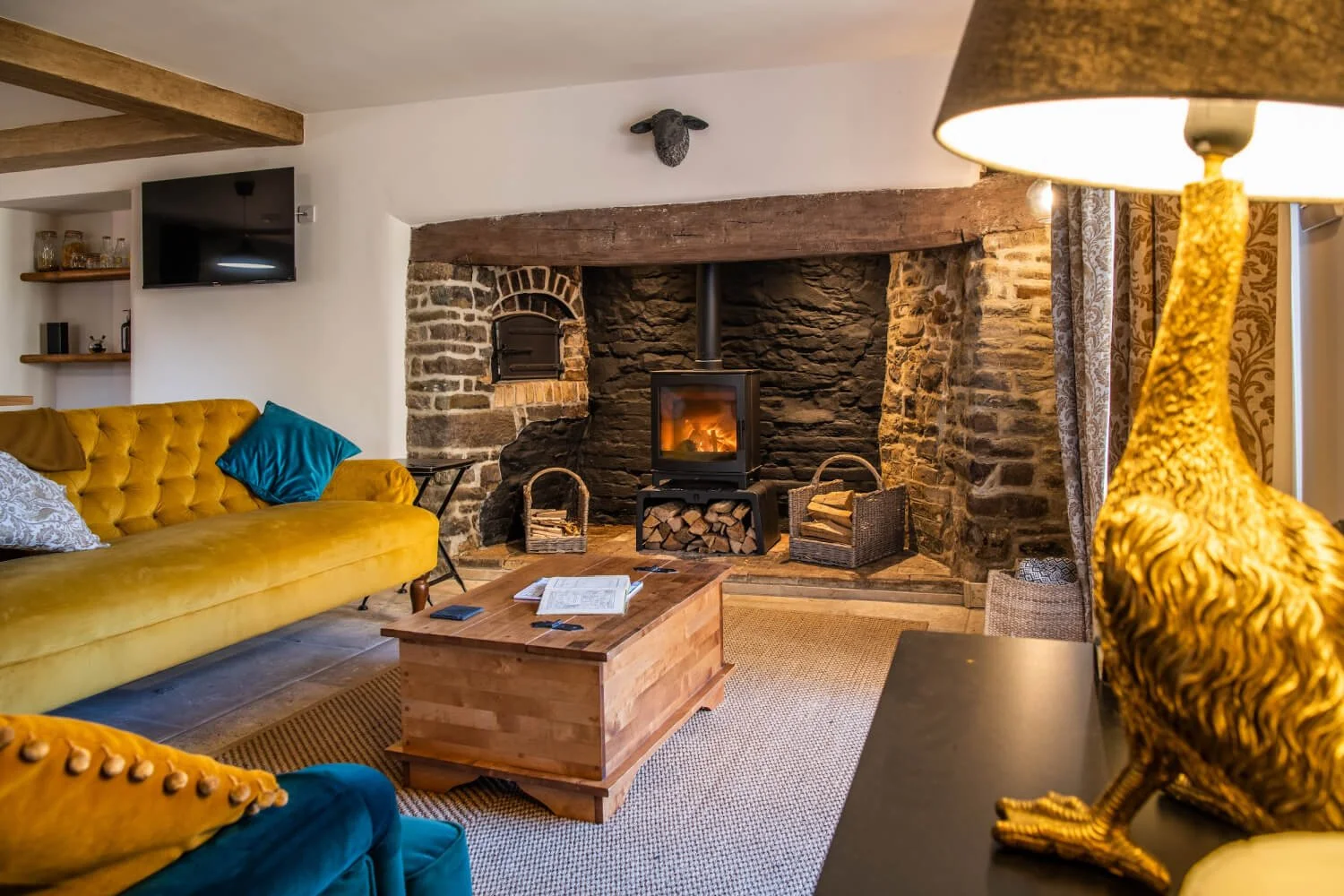Cozy living room with a stone fireplace, a wood-burning stove, yellow and blue furniture, and decorative accents.