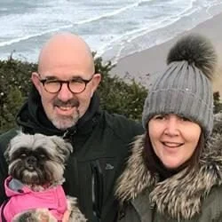 Sarah and David with their small dog Prudence on a beach with ocean waves in the background.