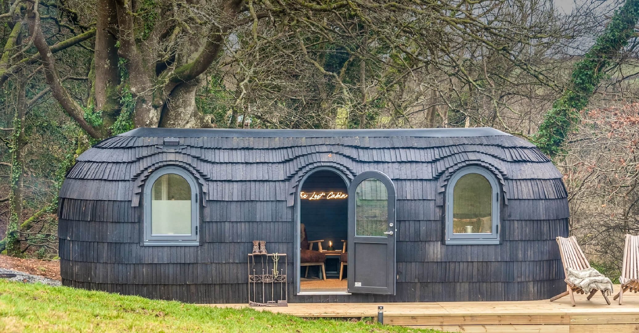 A small black cabin with arched windows in a wooded area, with outdoor chairs and a porch.