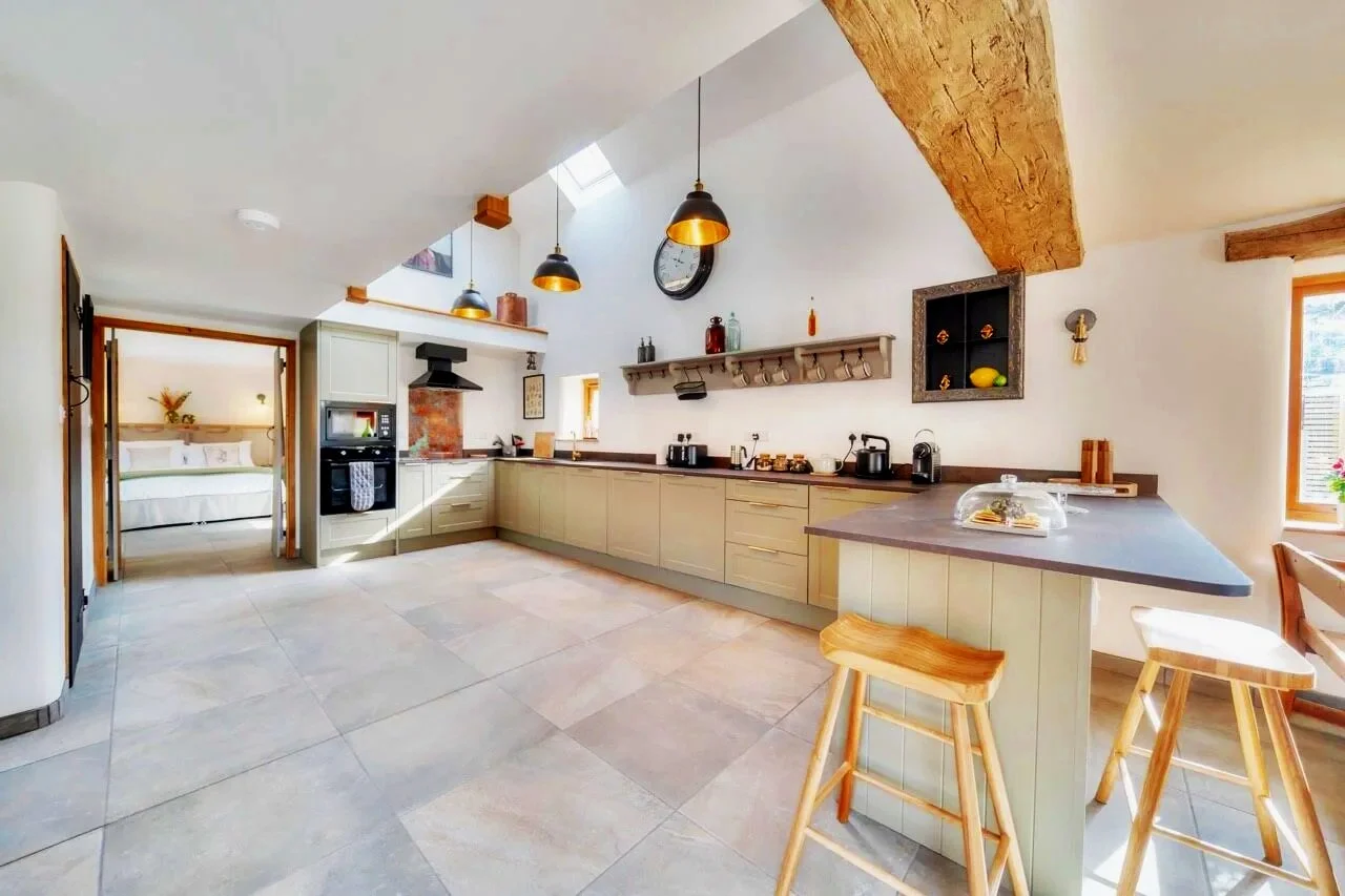 Modern open-concept kitchen with light gray cabinets, a large island, three bar stools, pendant lights, and a view into a bedroom.
