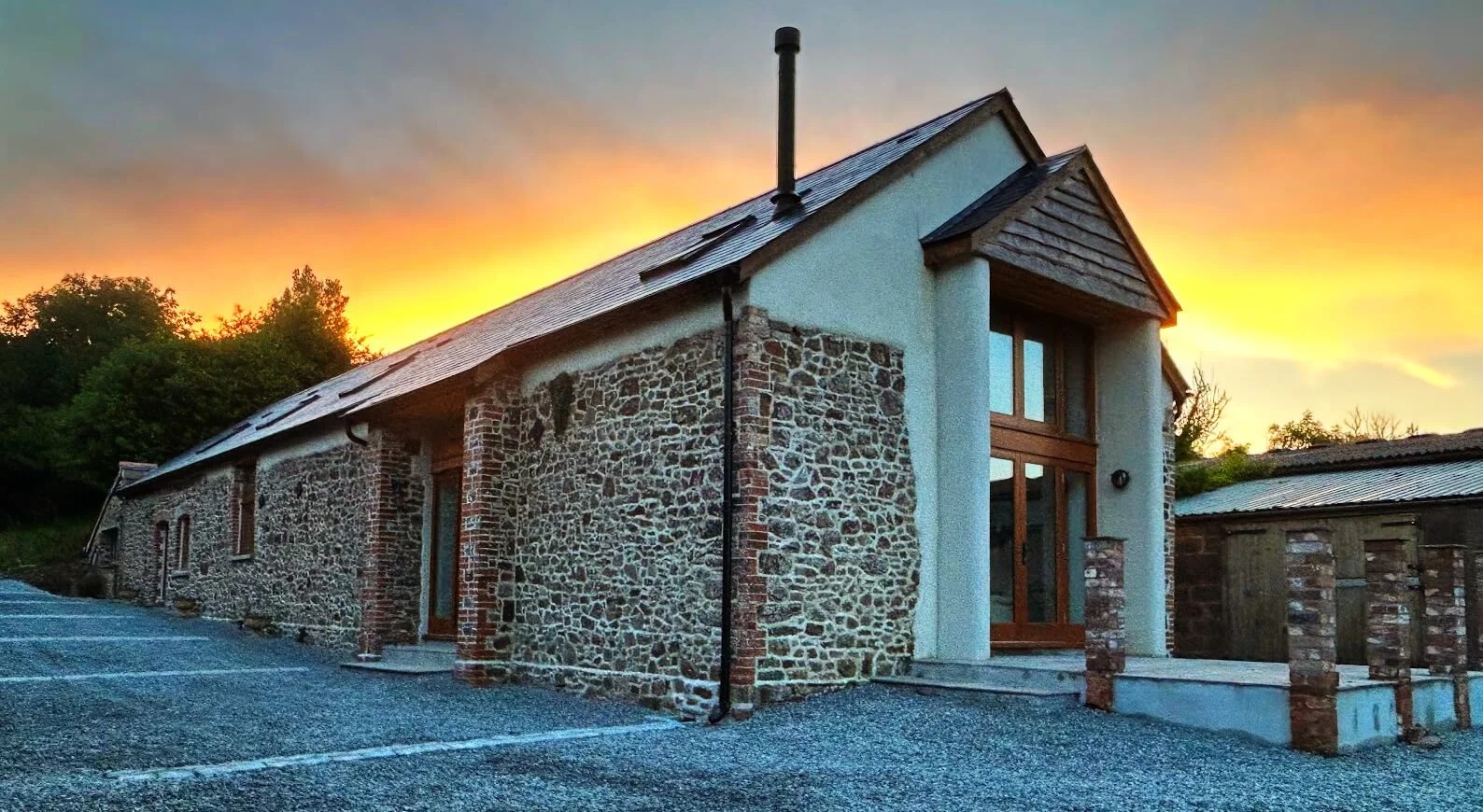 A rustic stone and stucco house during sunset with a parking lot in front and trees in the background.