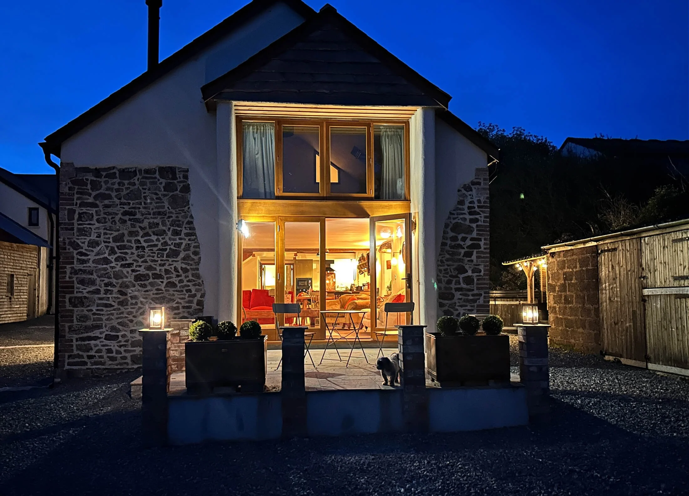 A two-story house with stone and white walls, lit from inside, with a small porch and outdoor furniture. A small dog is on the porch, and the house is surrounded by a gravel yard and wooden fences, during nighttime.