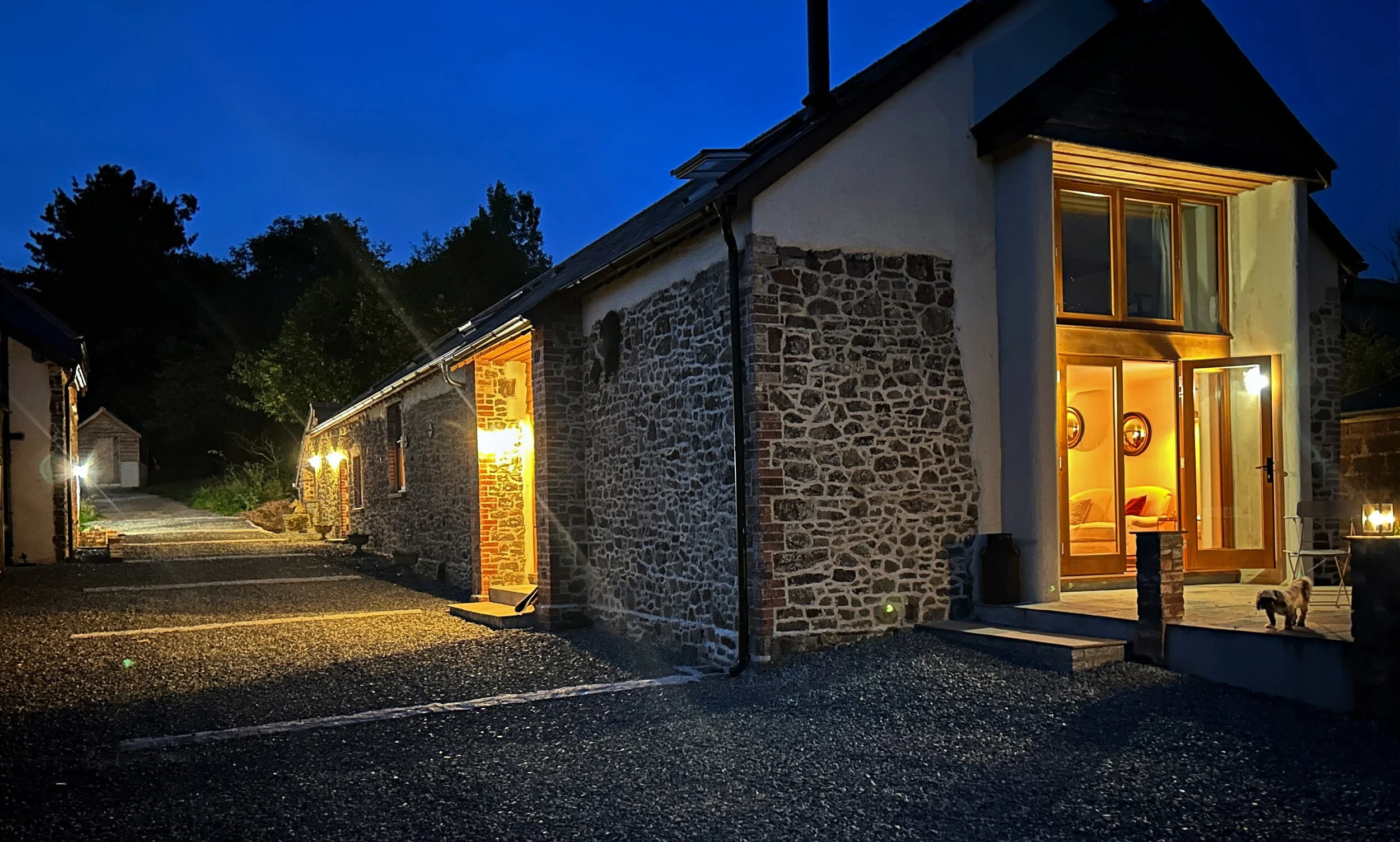 Nighttime view of a house with stone walls and large windows illuminated from inside, with a small dog on the porch. The driveway is empty with marked parking spaces.