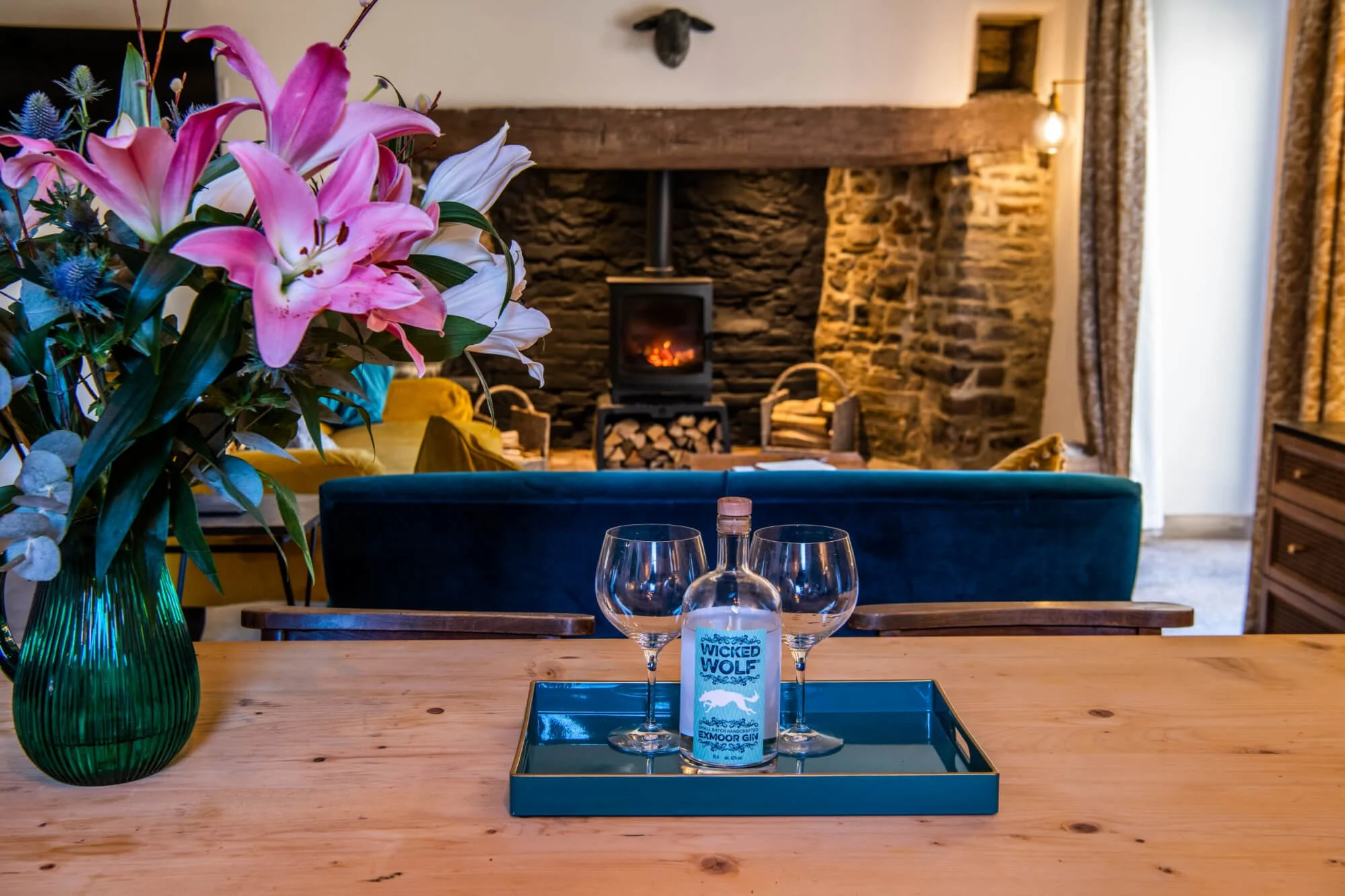 A wooden table with a green vase filled with pink and white lilies and other flowers, a tray with a bottle of Wicked Wolf Exmoor gin and two empty glasses, in a cozy living room with a stone fireplace, a fire burning inside, yellow and teal seating, 