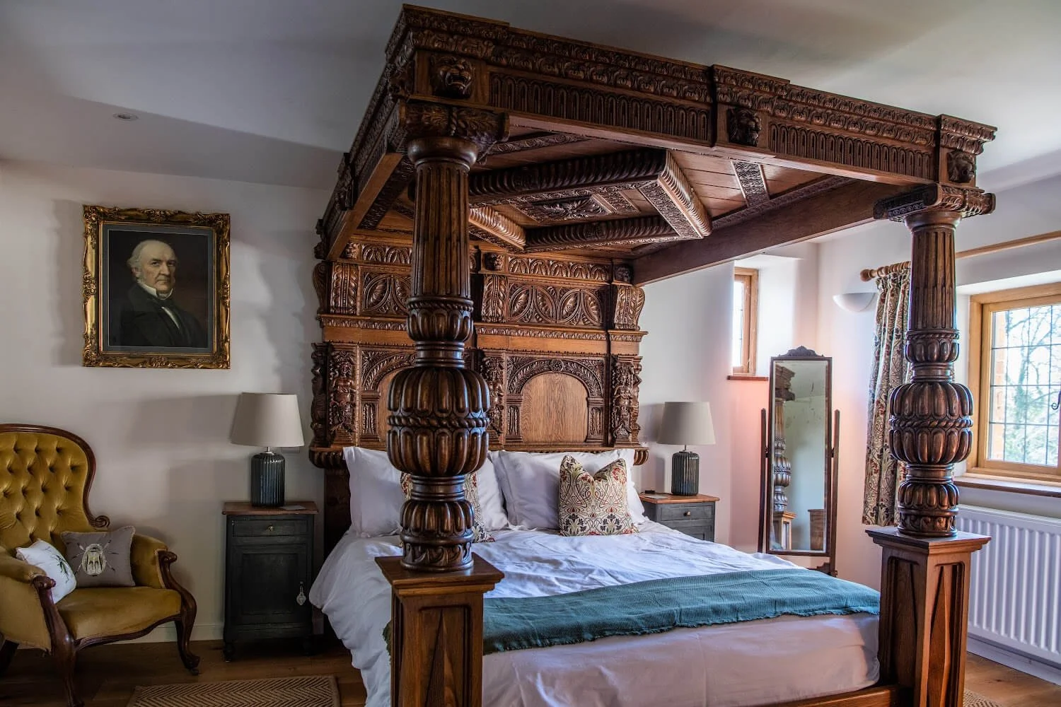 A bedroom with a large ornate wooden four-poster bed with carved details, white bedding, and a decorative pillow. To the left, there is a yellow tufted armchair with pillows, and next to it is a small dark nightstand with a lamp. On the wall above th