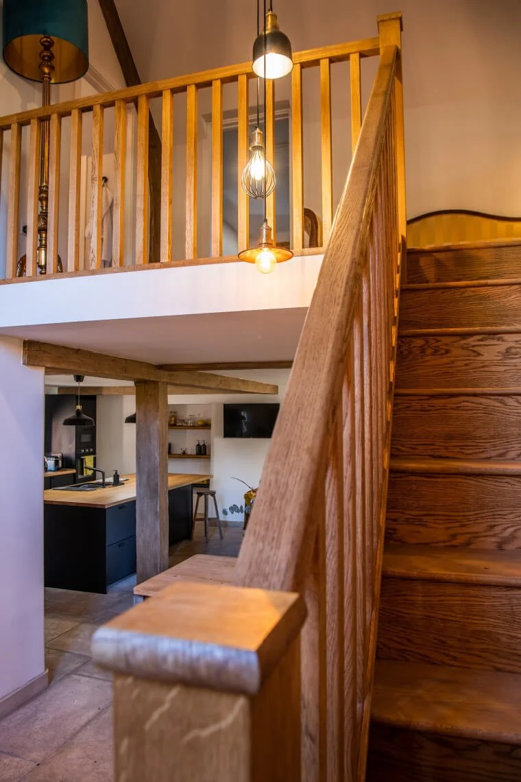 Interior view of a wooden staircase leading to a loft area with hanging pendant lights, overlooking a kitchen with black cabinets and a wooden countertop.