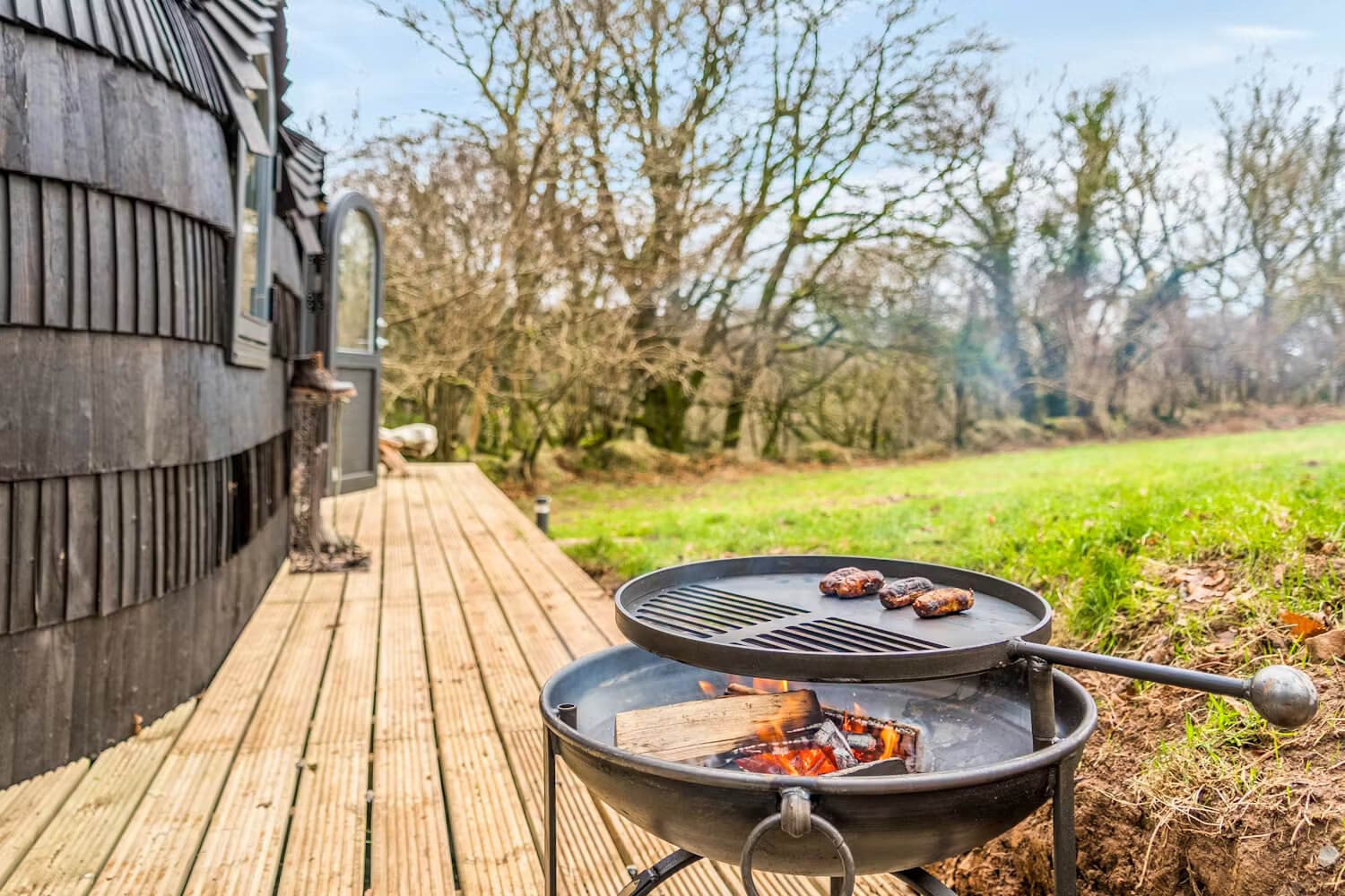 A charcoal grill on a wooden deck with food cooking on it, next to a house with dark wooden siding, in a backyard with grass and leafless trees in the background.