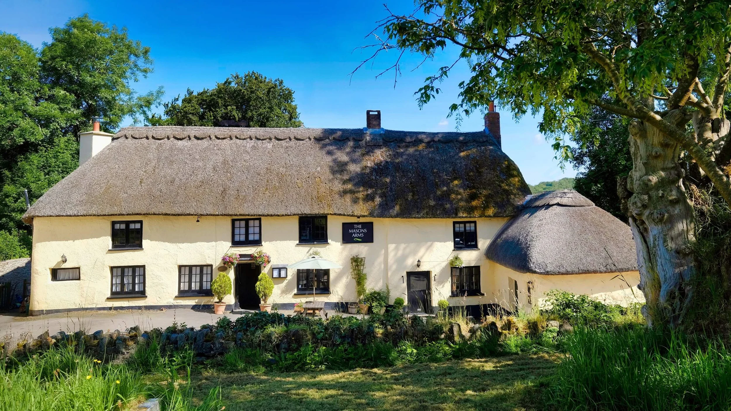 Thatched-roof cottage with cream-colored walls, multiple windows with black frames, a small garden in front with potted plants, and a sign reading 'The Masons Arms' on the wall, surrounded by trees and greenery.