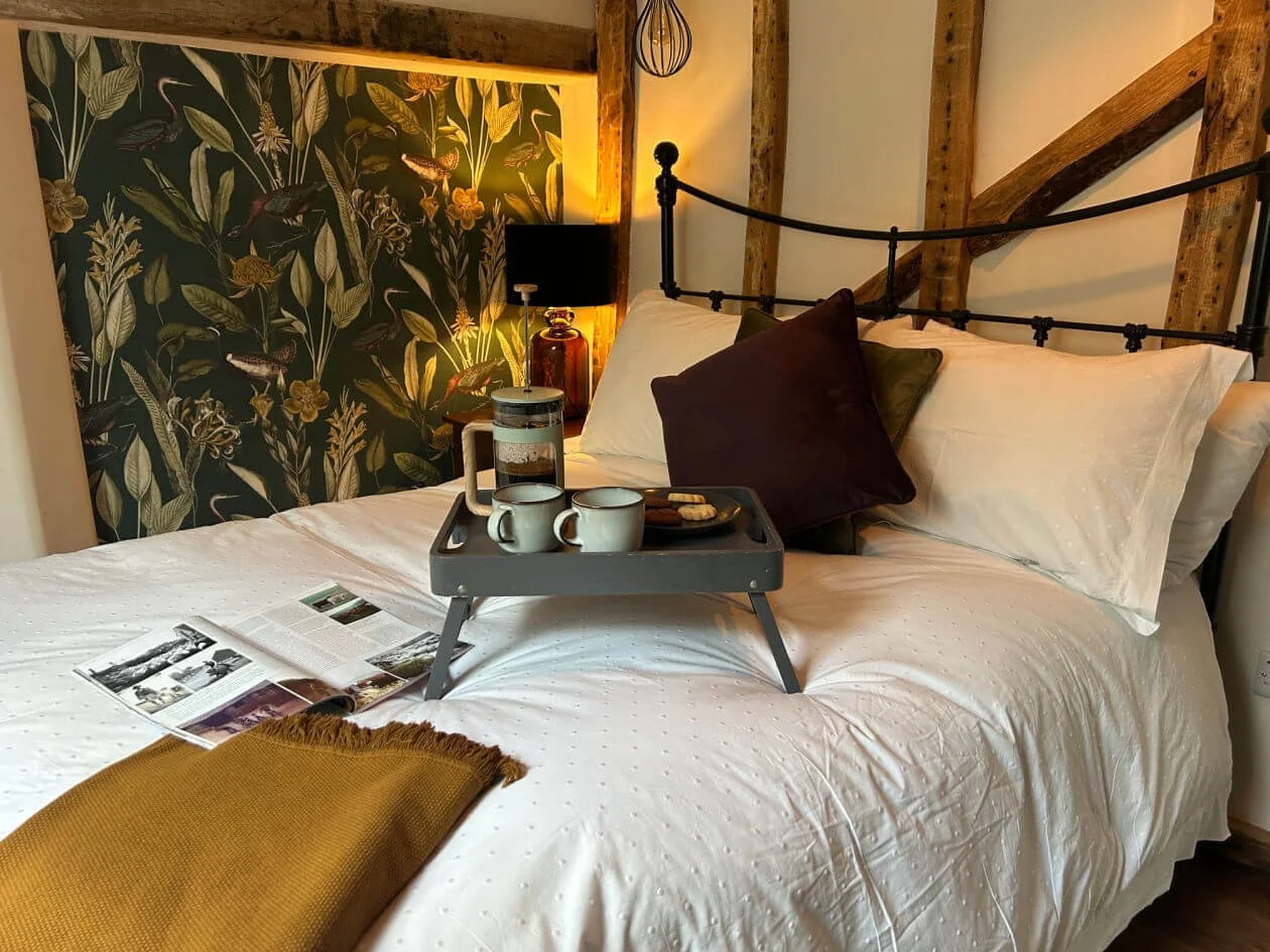 A cozy bedroom with a neatly made bed, decorative pillows, a tray with coffee cups and snacks, an open magazine, and a mustard-colored blanket. The wall behind the bed has a black floral wallpaper with plants, and there is a black bedside lamp with a