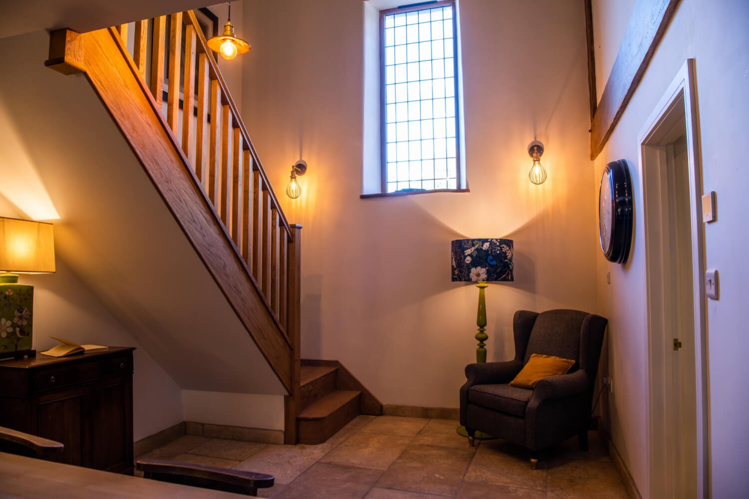 Interior of a cozy living space with a staircase, tall window, armchair, floor lamp, wall clock, side table, and decorative lights.