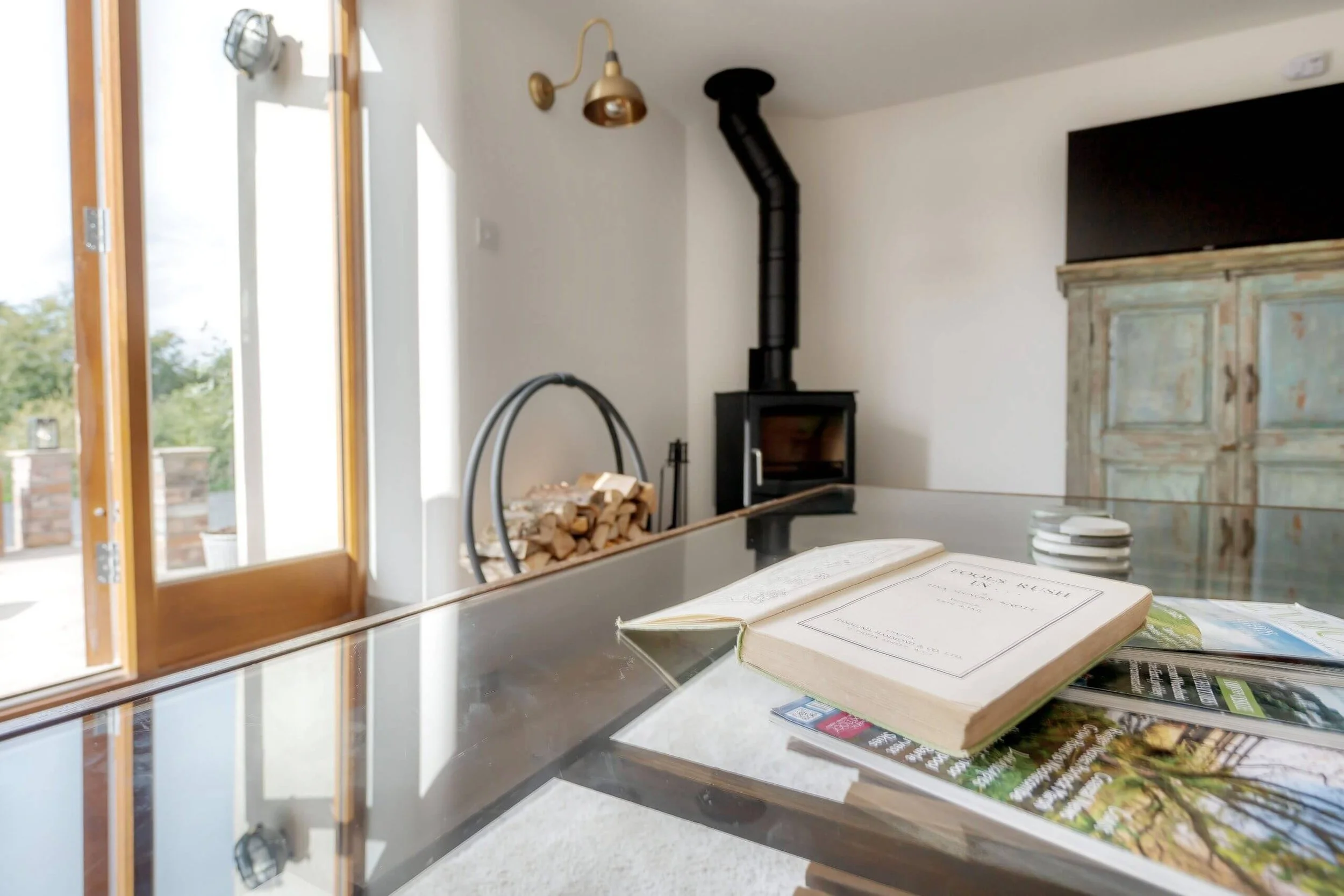 Close-up of an open book and magazines on a glass table in a cozy living room with a wood stove, a firewood rack, a rustic cabinet, and a large window with natural light.