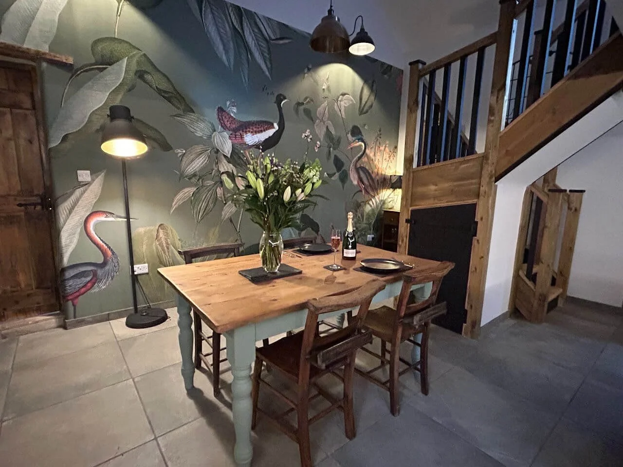 A dining area with a wooden table set for two, featuring black plates, a champagne bottle, and two glasses of champagne. The table is decorated with a tall vase of white lilies. The background has a wall with a colorful bird and foliage mural, and th