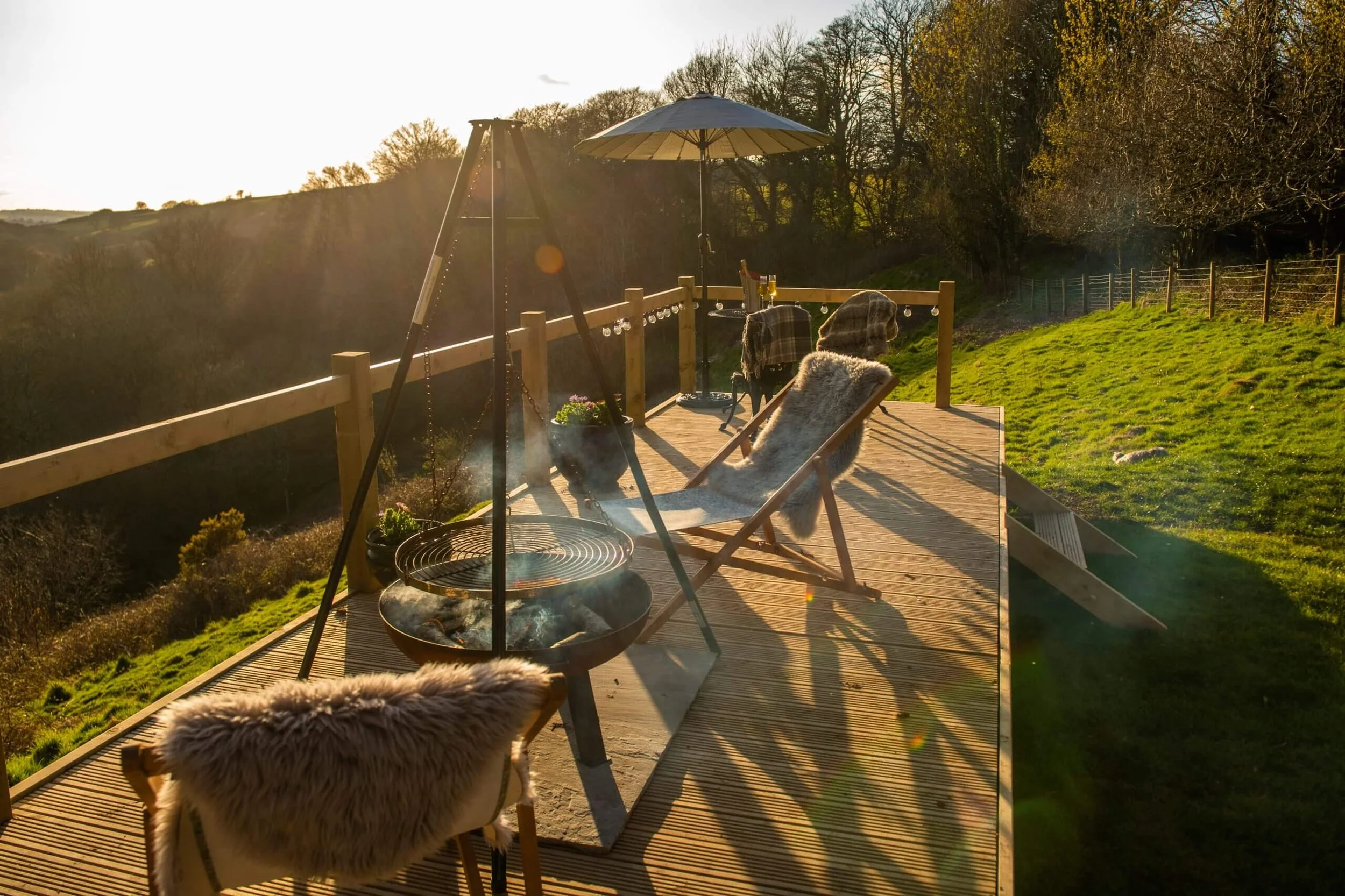 Sunlit outdoor wooden deck with two chairs, an umbrella, a fire pit, and a view of the hilly landscape with trees in the background during sunset.