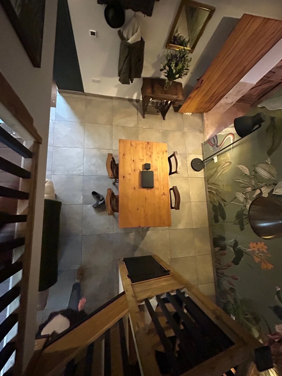 From a high angle, the photo shows a dining area with a wooden table and four chairs, two chairs on each side. A black rectangular object and a small black item are on the table. The floor has beige tiles. To the right, there's a floor lamp with a bl
