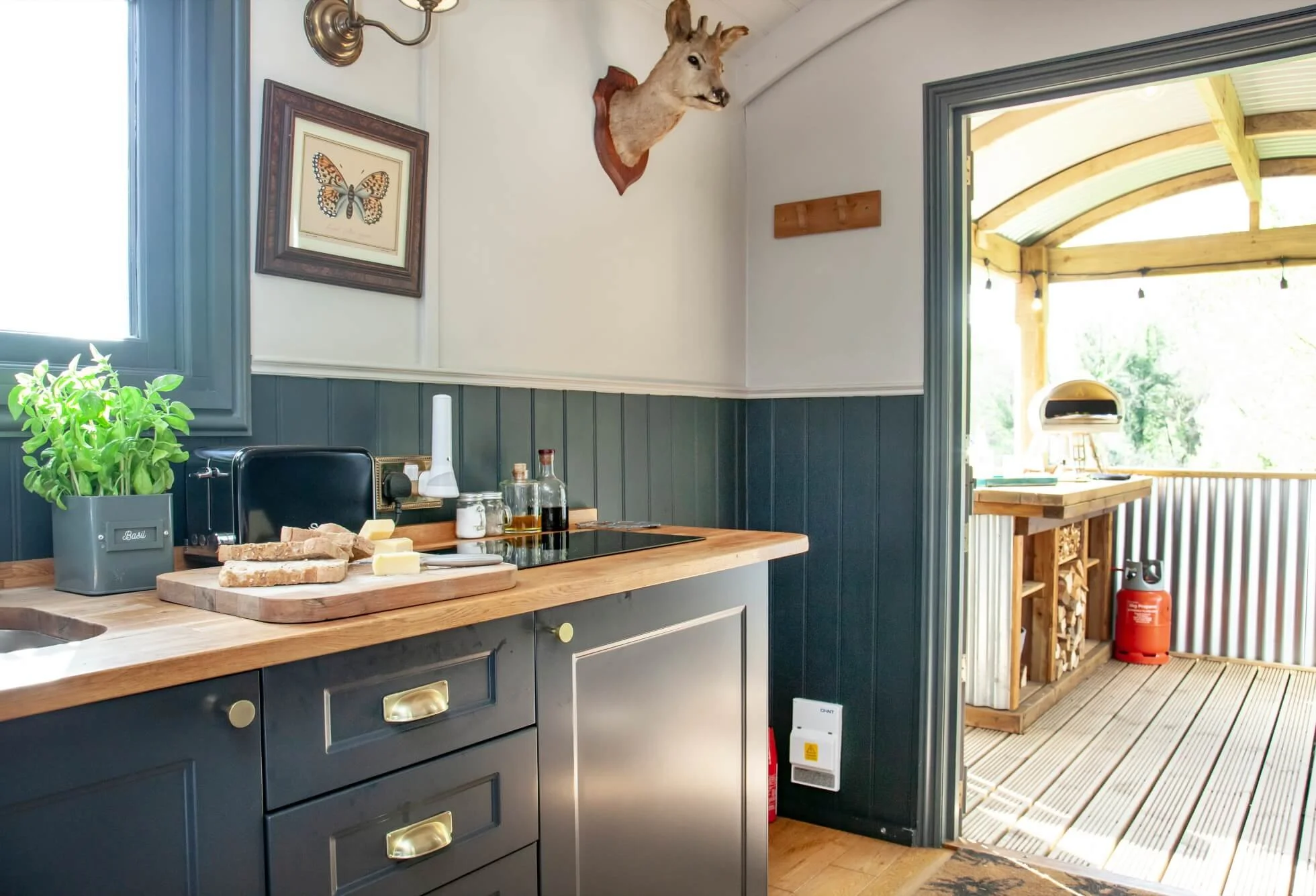 A kitchen with a black cabinet and wooden countertop, plants, cheese, bread, oil bottles, framed butterfly art, a mounted deer head, and an outdoor covered porch with a wooden countertop, fire extinguisher, and wood storage.