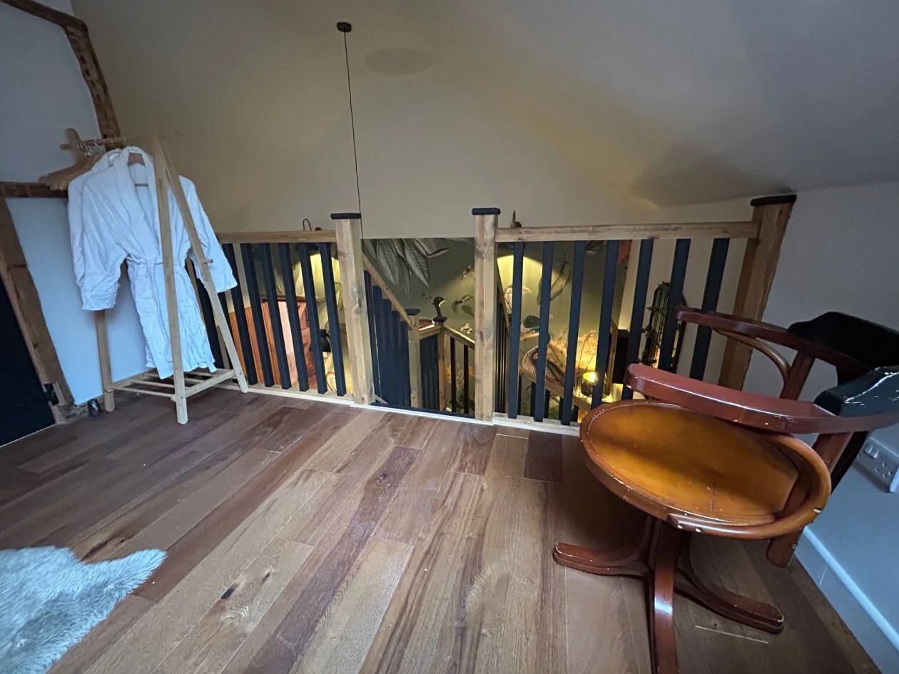 A cozy loft area with wooden flooring, a wooden rocking chair, and a clothes rack with a white bathrobe hanging on it. There is a staircase leading down to a decorated lower level with a mural.