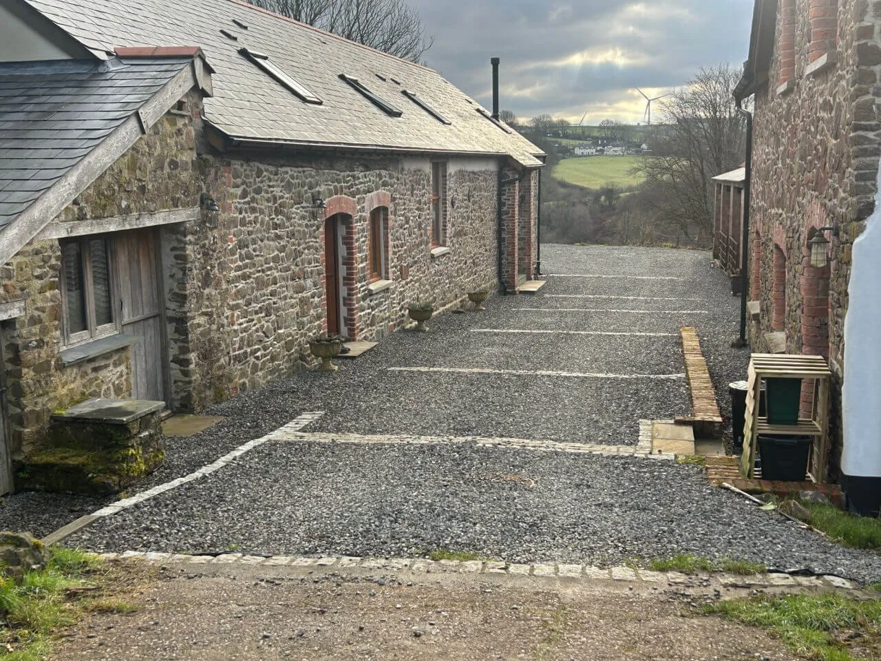 A gravel parking area between two stone buildings with small plants in planters along the walls, and a scenic countryside view with wind turbines and rolling hills in the distance.